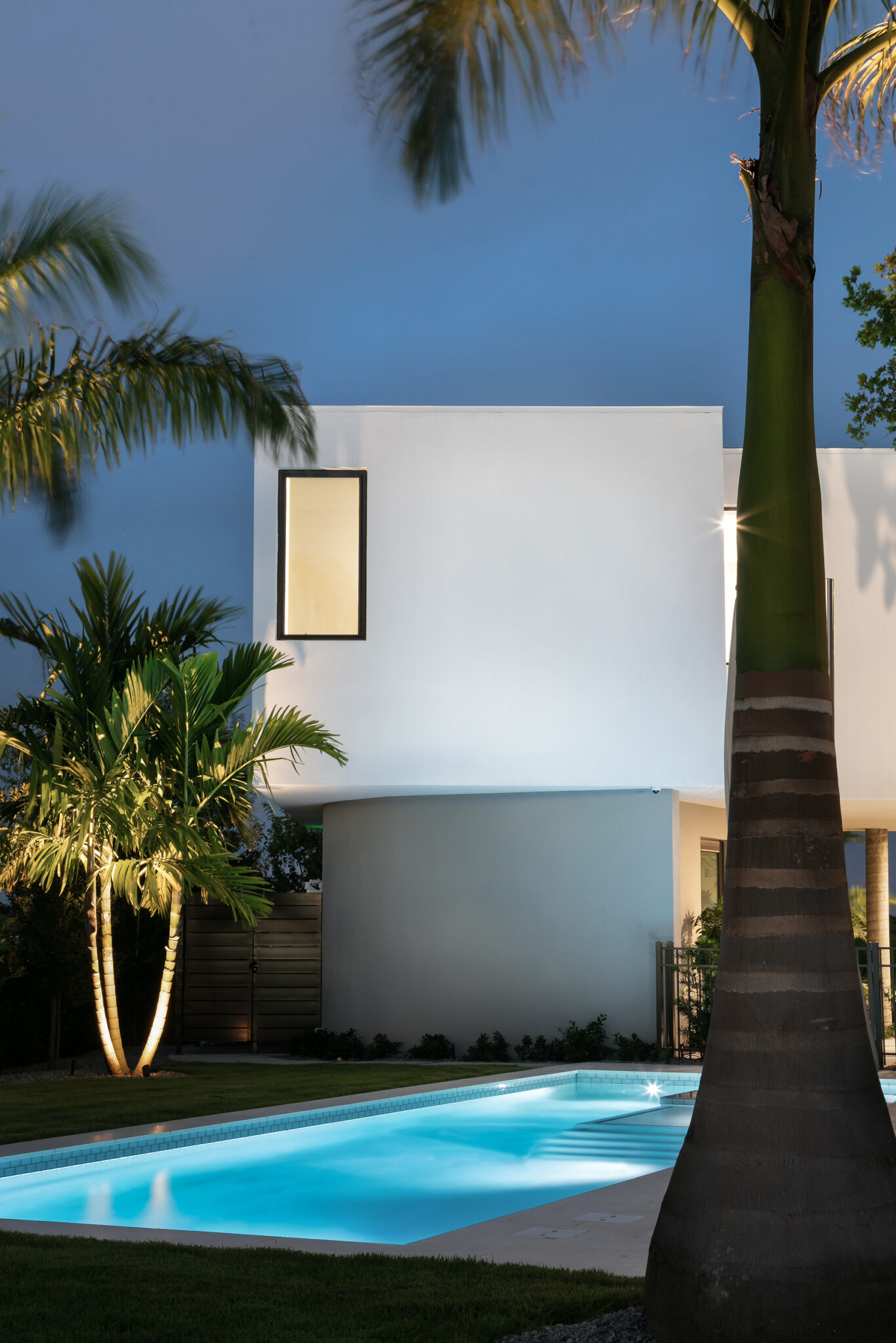 Pool lit at night beside a white cube volume and palms