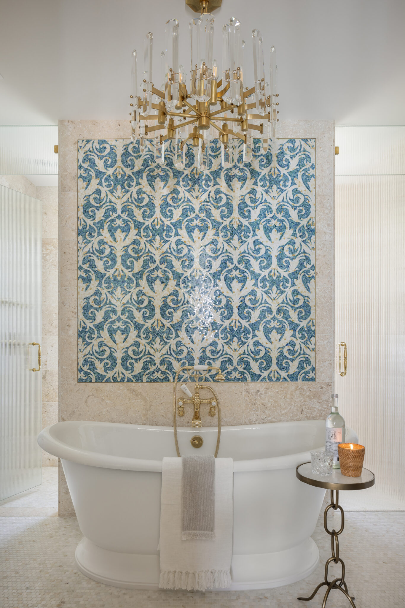Freestanding tub under a blue patterned tile wall and gold chandelier