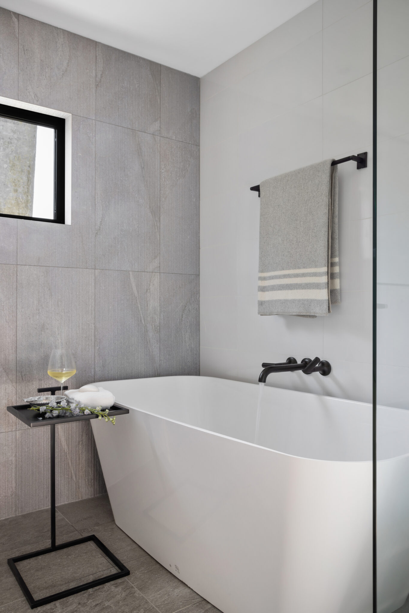 Freestanding tub in a gray tile bath with a black-framed window