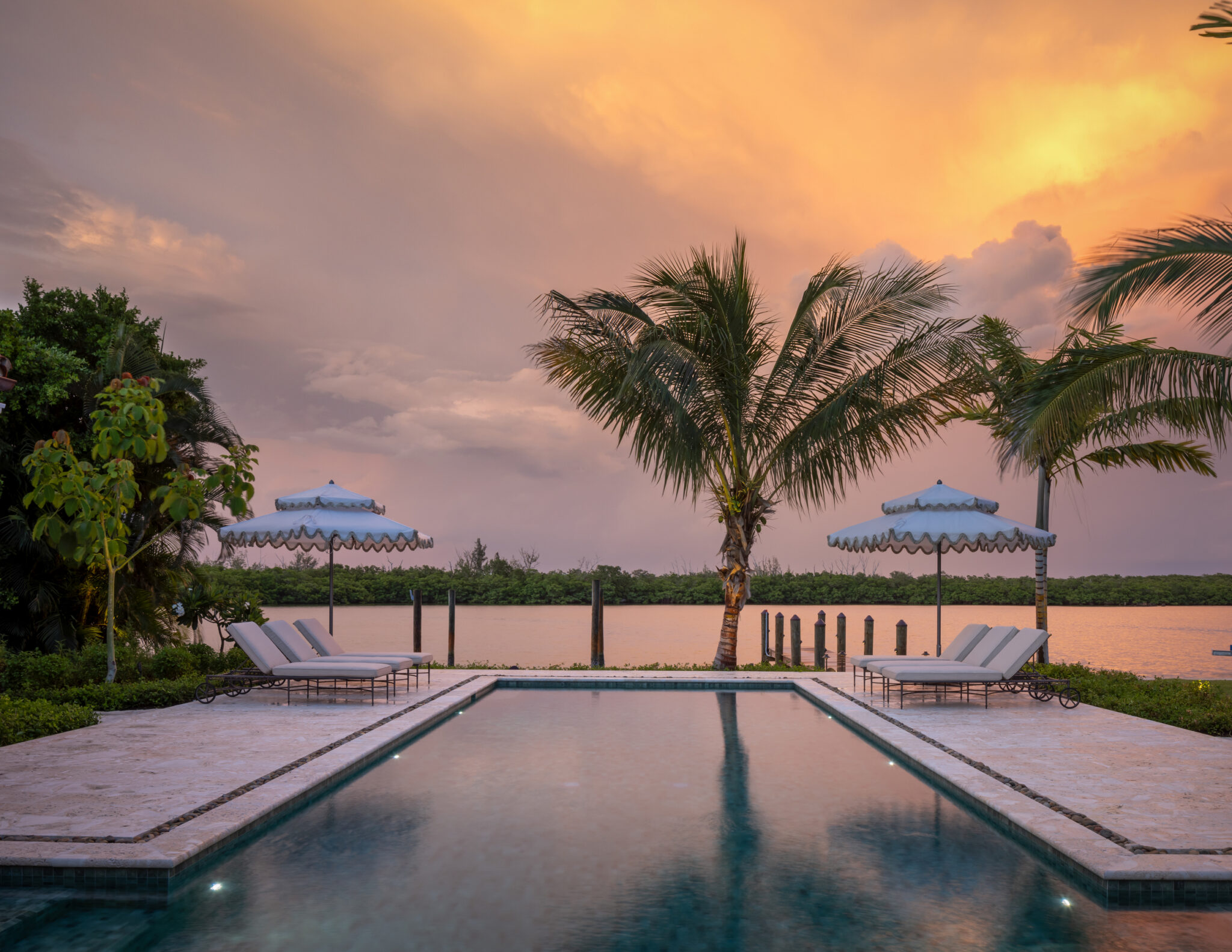 Pool reflecting a sunset sky, with palms and umbrellas at the water's edge