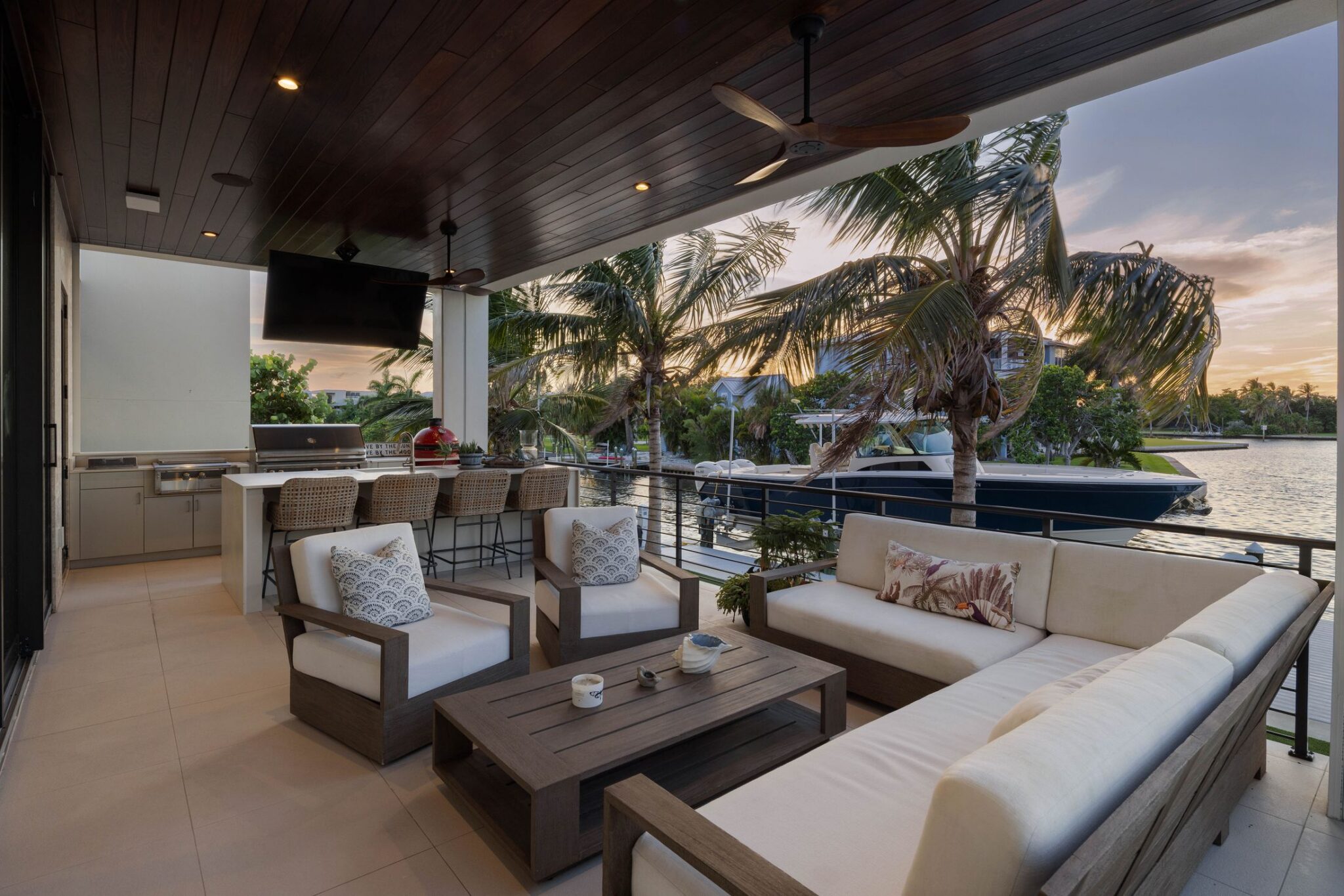 Covered outdoor living room with an outdoor kitchen and bar seating, facing the bay at sunset