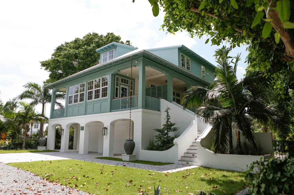 An elevated coastal cottage with sea-green trim with a large porch, multiple windows, stairs leading up to the entrance, and surrounded by palm trees and greenery