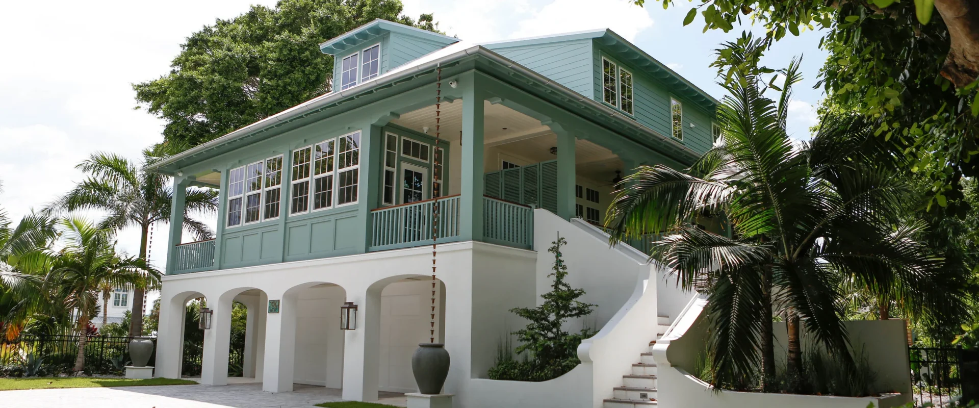 An elevated coastal cottage with sea-green trim with a large porch, multiple windows, stairs leading up to the entrance, and surrounded by palm trees and greenery