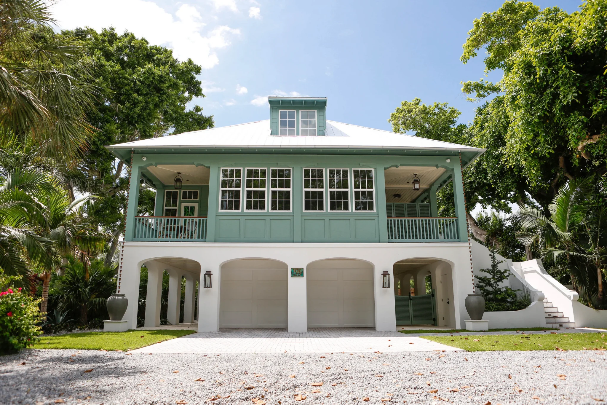 An elevated cottage with sea-green trim, a metal roof, large front windows, and a covered porch, surrounded by lush trees and greenery