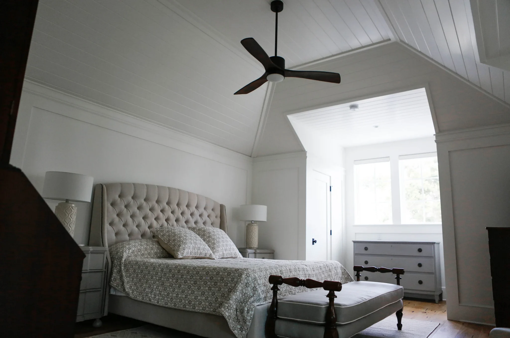 Bright bedroom with white walls and ceiling, featuring a beige tufted headboard, patterned bedding, nightstands with lamps, a ceiling fan