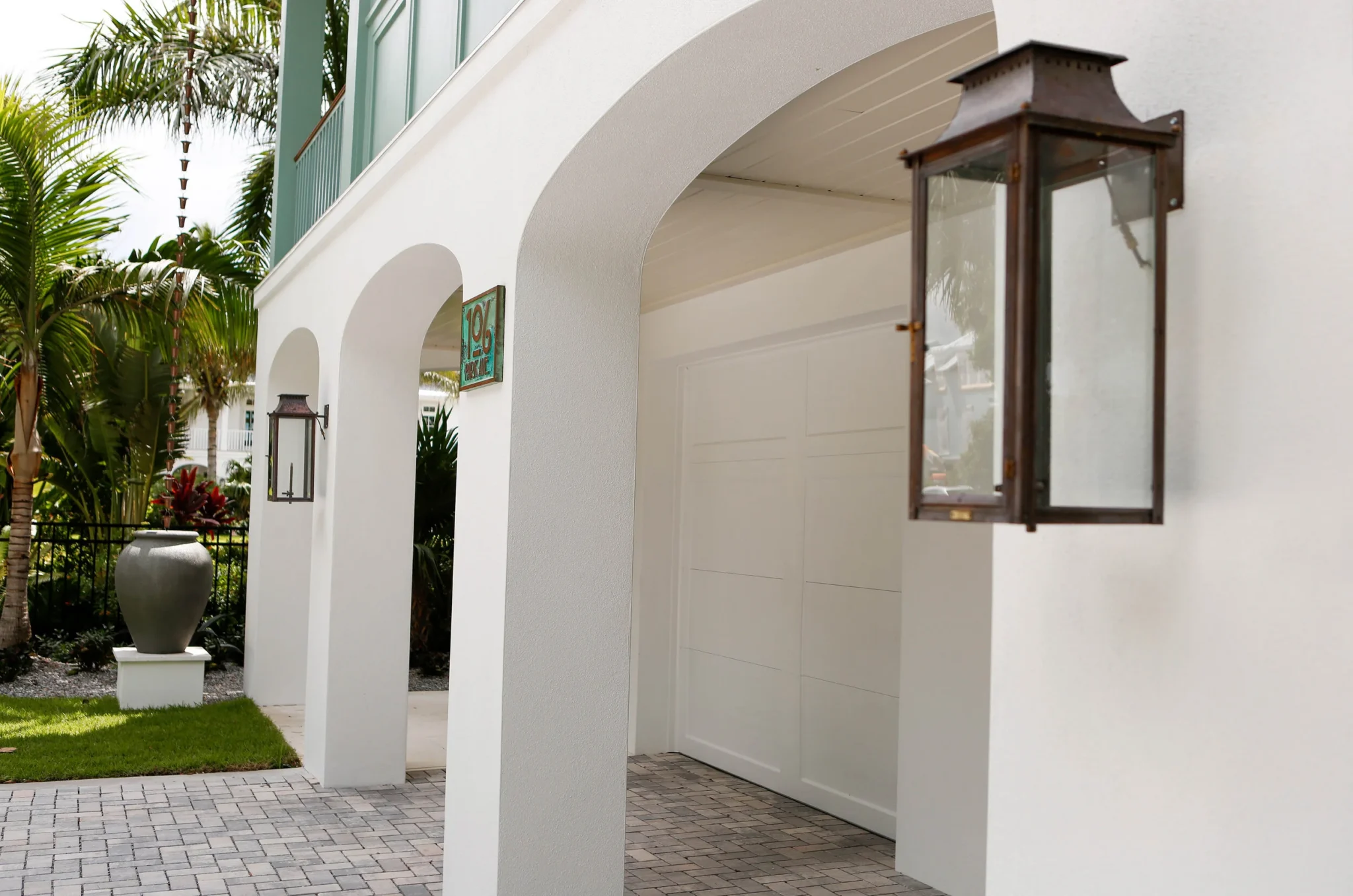 Exterior of a modern white house with arched entryways, black lantern-style wall lights, a decorative sign, a large gray urn on a pedestal