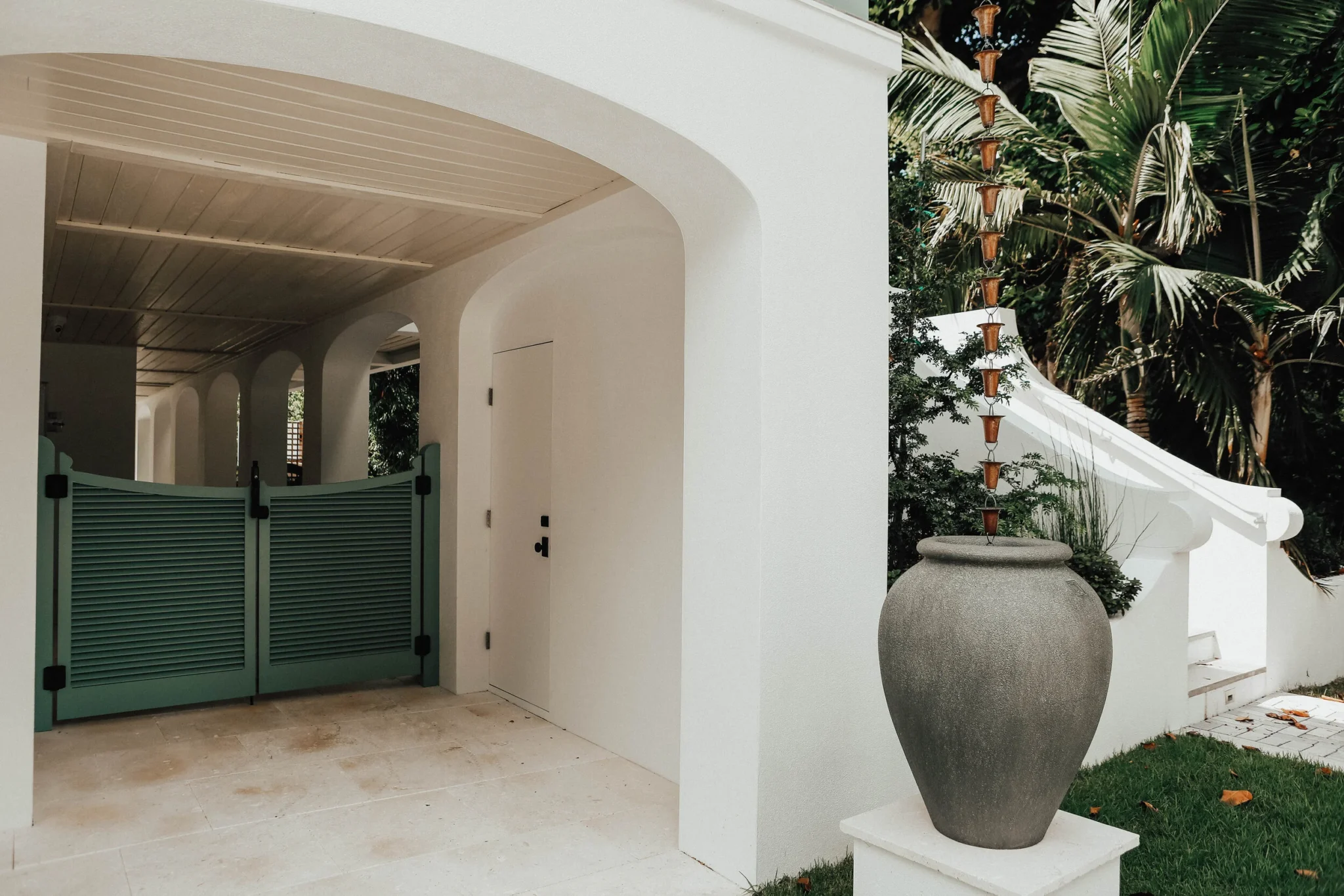 Exterior of a white modern house with an arched entryway, green gate, and a large outdoor ceramic pot with a tall plant and hanging decorative cones