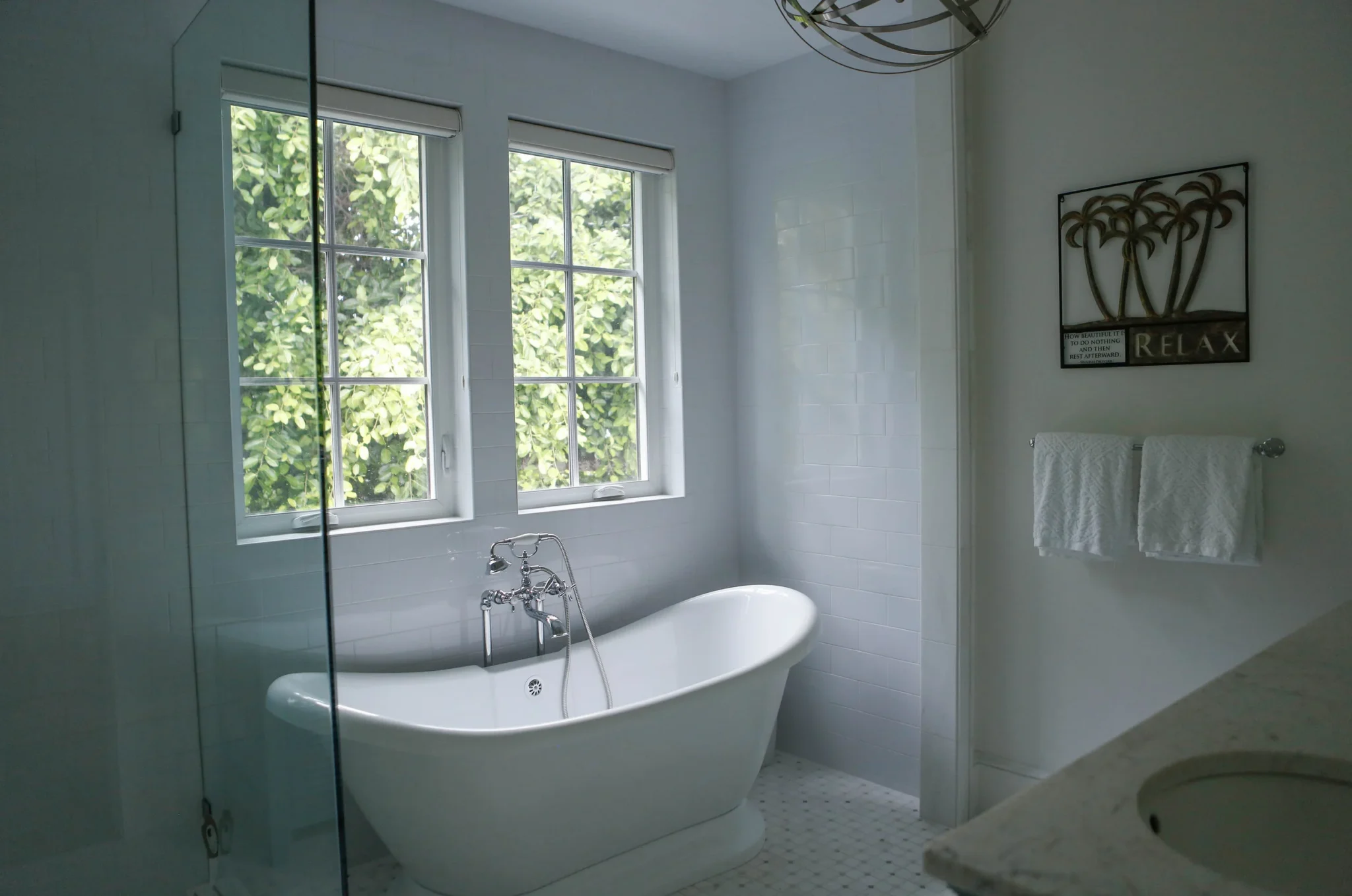 Bathroom with a white clawfoot bathtub, two windows with greenery outside, white tiled wall, towels on a towel rack, wall art