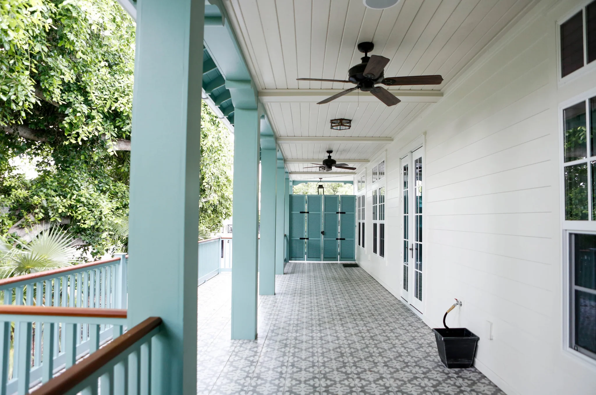 Covered balcony with patterned tile floor, white siding walls, multiple windows, teal support columns, teal shutters at the end, ceiling fans