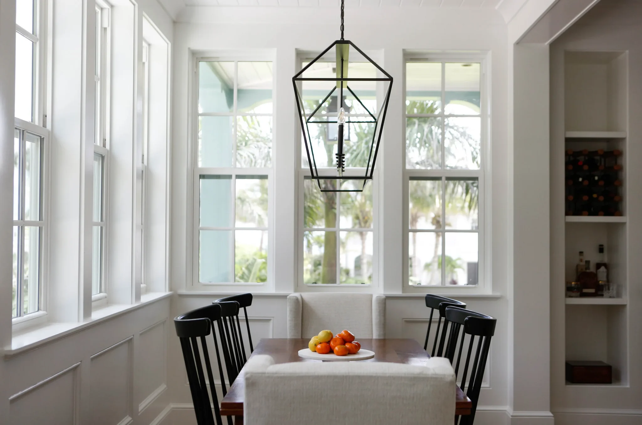 Bright dining room with large windows, a wooden table with a bowl of fruit, black chairs, and a geometric pendant light