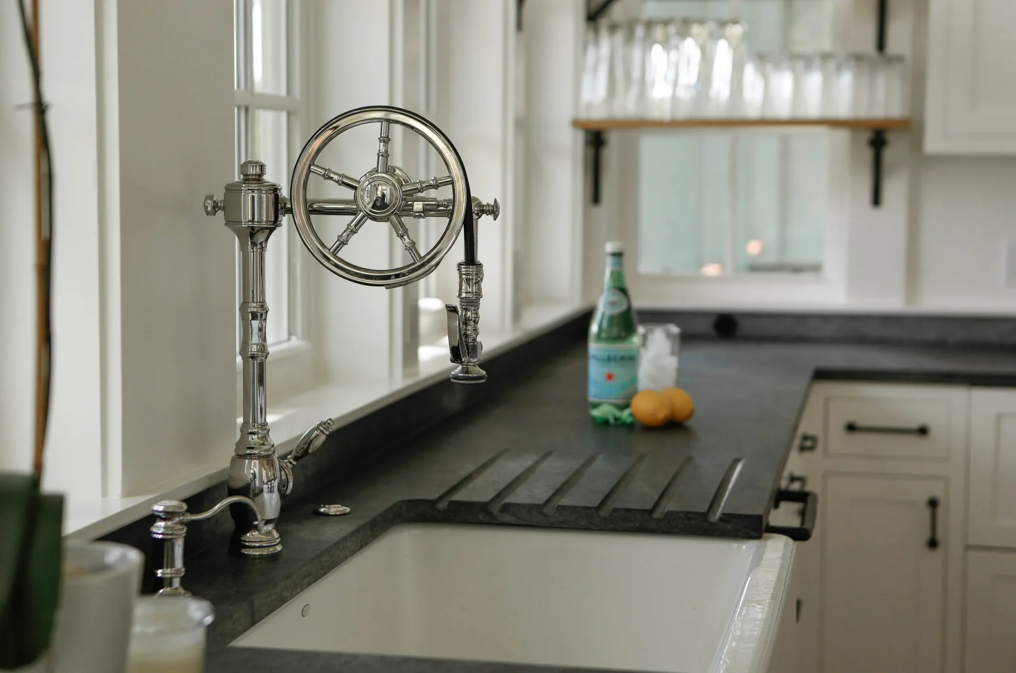 Kitchen sink with a vintage chrome faucet in front of a window, with a bottle of sparkling water, a glass, and two lemons on the black countertop