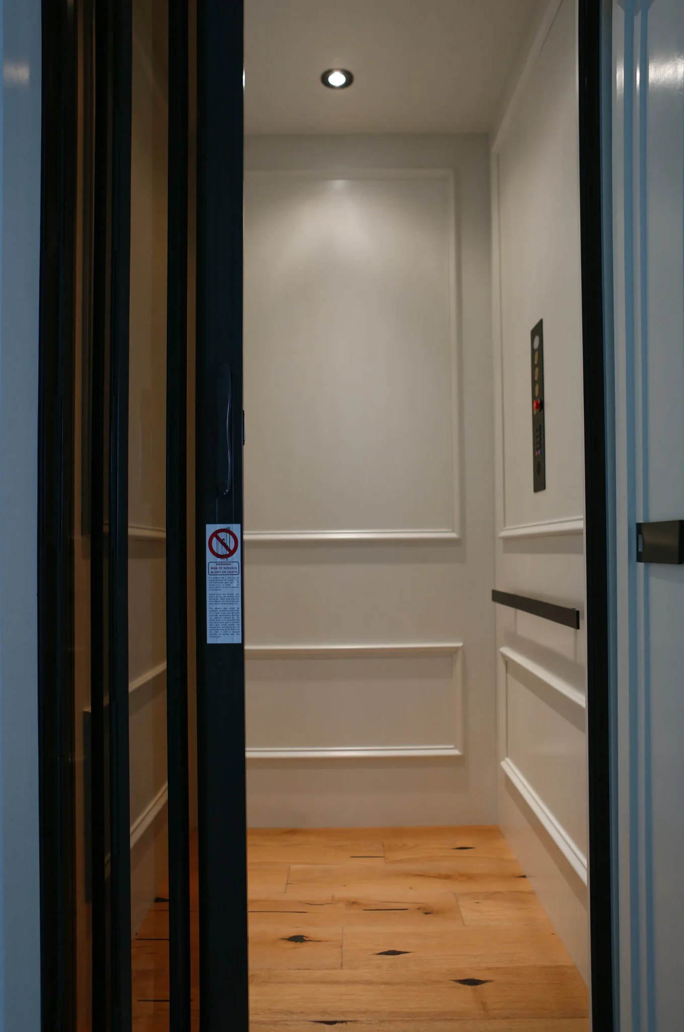 Inside a modern elevator with white paneled walls, a wood floor, and a ceiling light