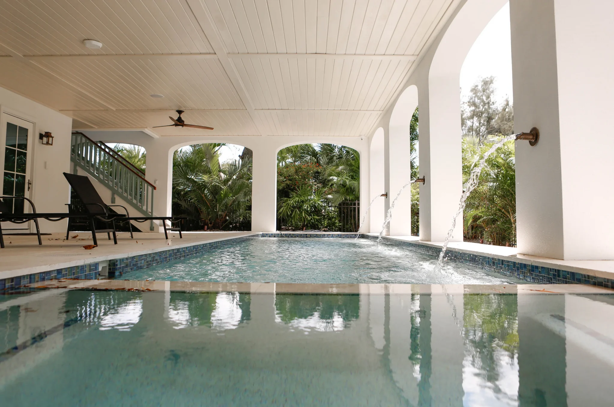 Indoor pool area with water features, lounge chairs, and lush greenery outside, under a ceiling with ceiling fan and arched openings