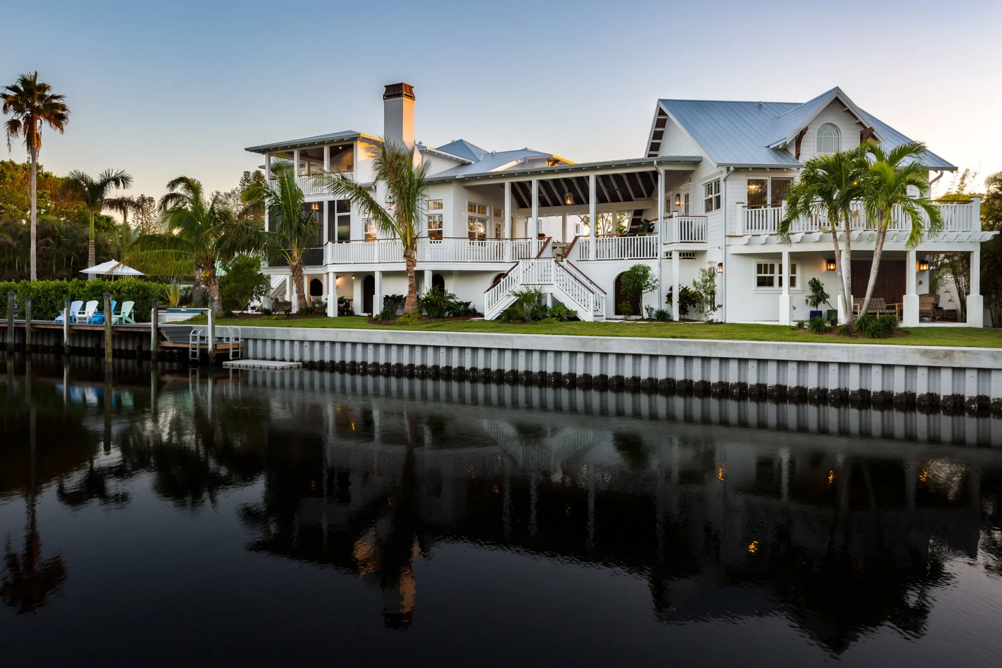 A large white house by a waterway with palm trees, a deck, and outdoor seating, reflected in the water