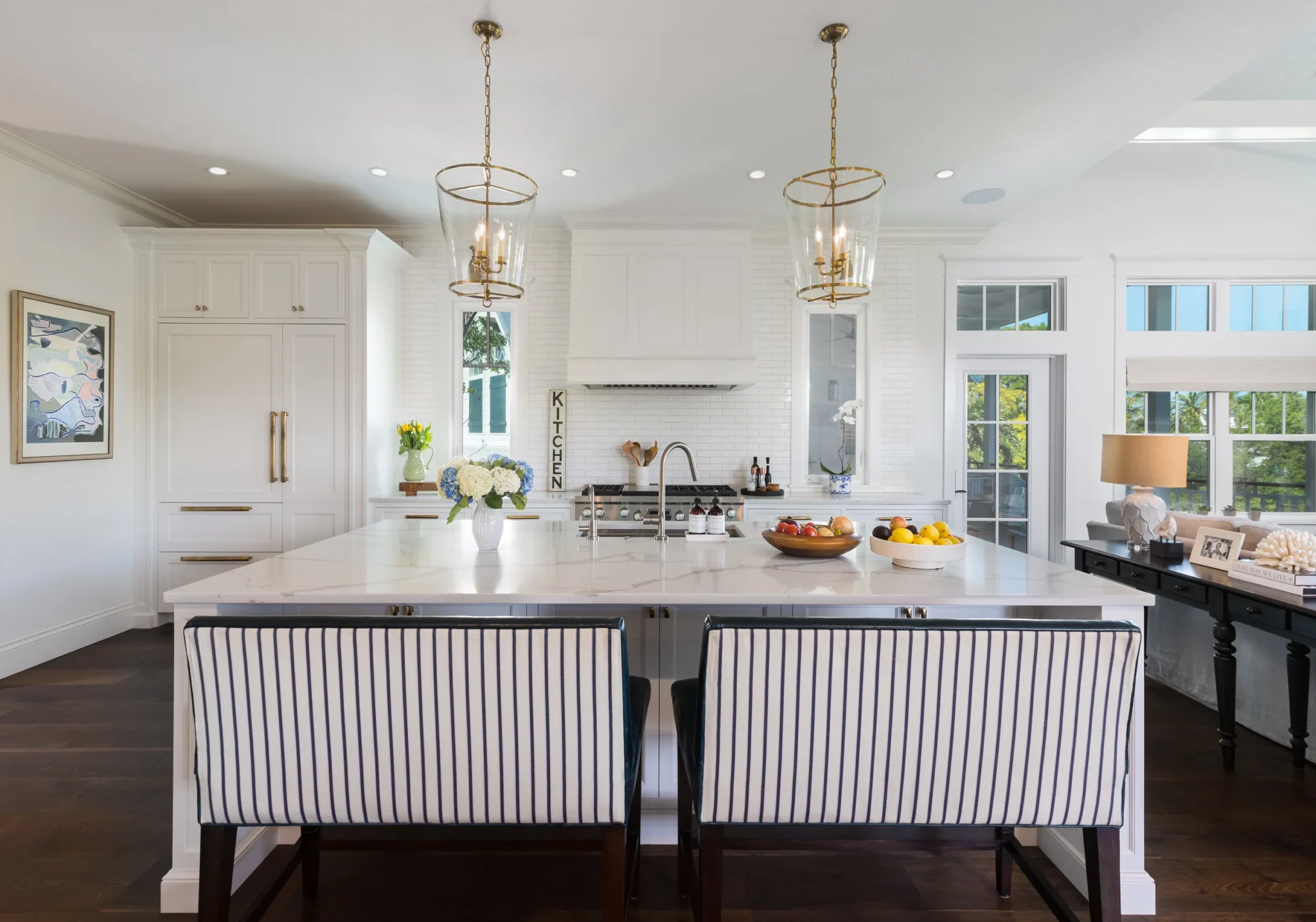 A bright kitchen with white cabinets, a large white island with a marble countertop, pendant lights, and dark hardwood floors