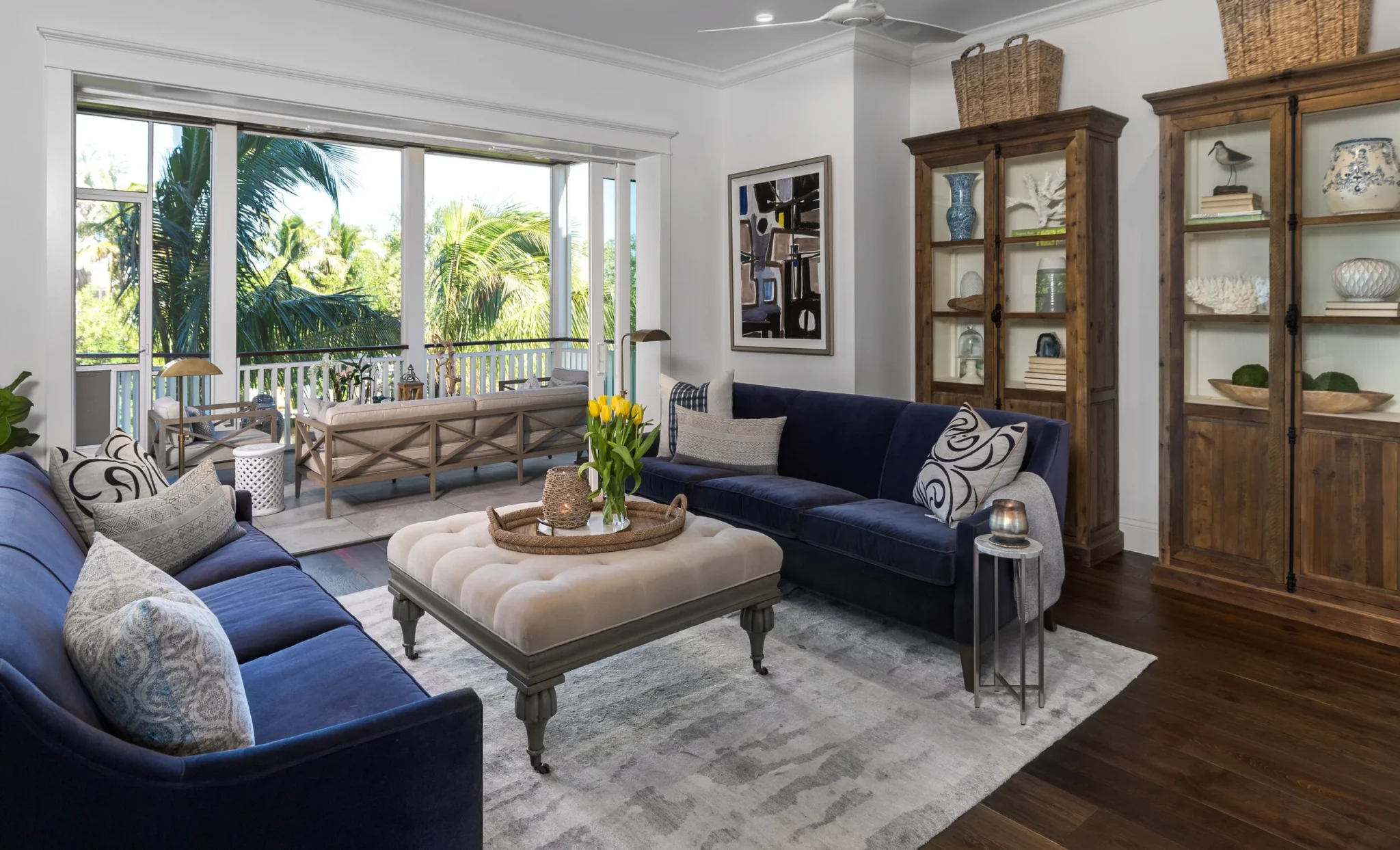 Living room with navy blue and gray sofas, a beige tufted ottoman with a tray of yellow tulips, featuring large windows overlooking greenery