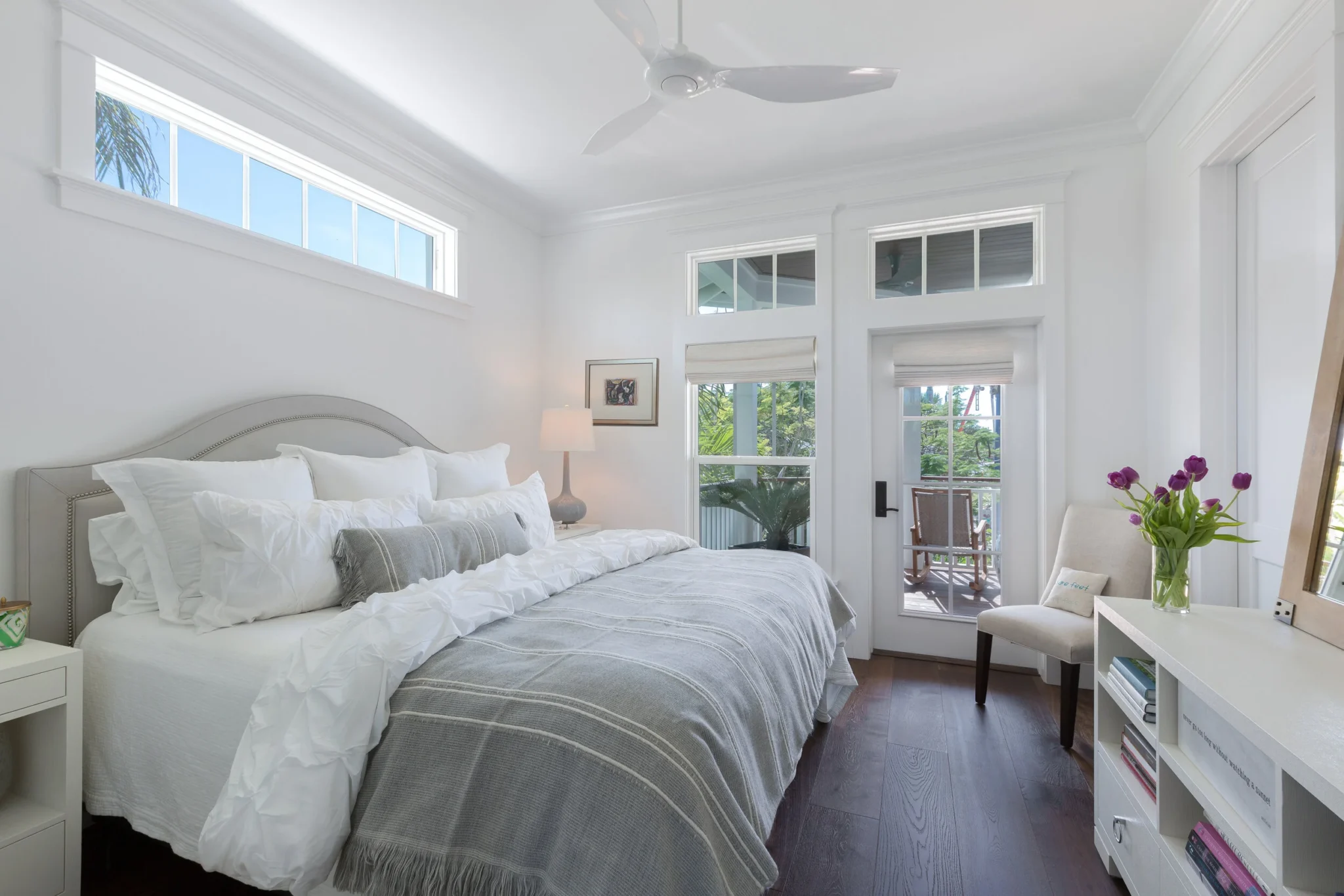 Bright bedroom with white walls, large window, and a balcony door, furnished with a bed, chair with flowers, bookshelf, and ceiling fan