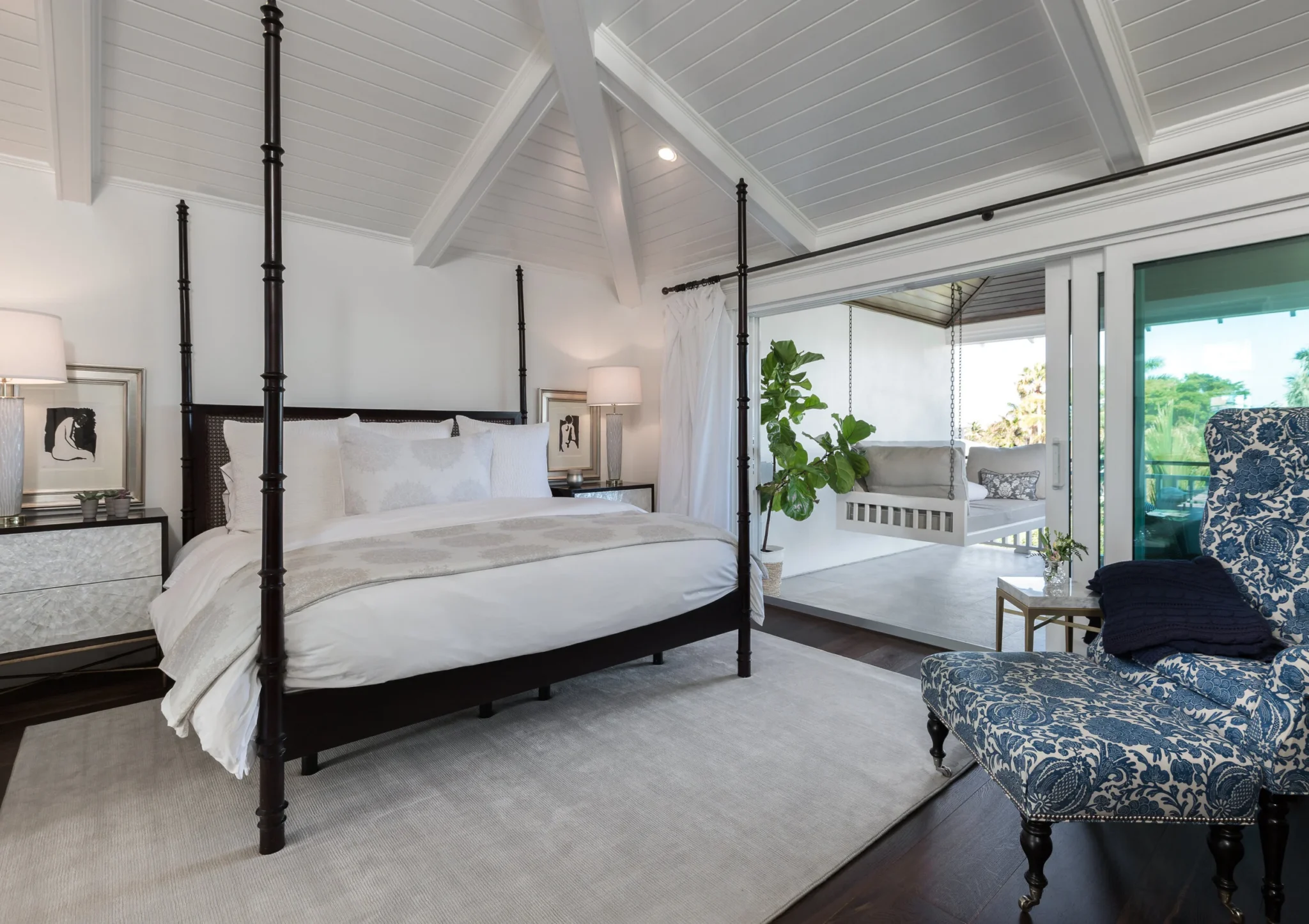 A bedroom with a four-poster bed, nightstands, lamps, and artwork, with a balcony featuring a hanging porch swing, potted plants, and outdoor seating