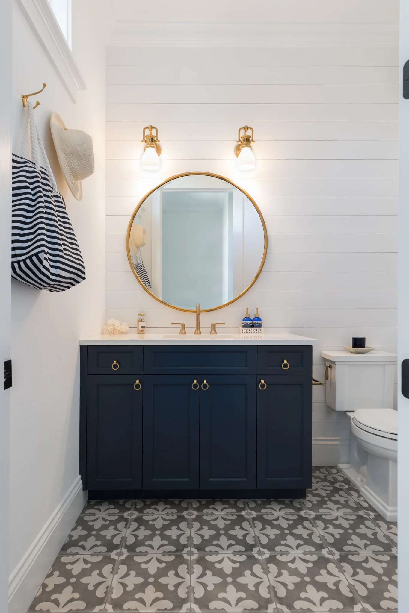 Bathroom with navy blue vanity, round mirror, white walls, decorative patterned floor tiles, and a toilet on the right