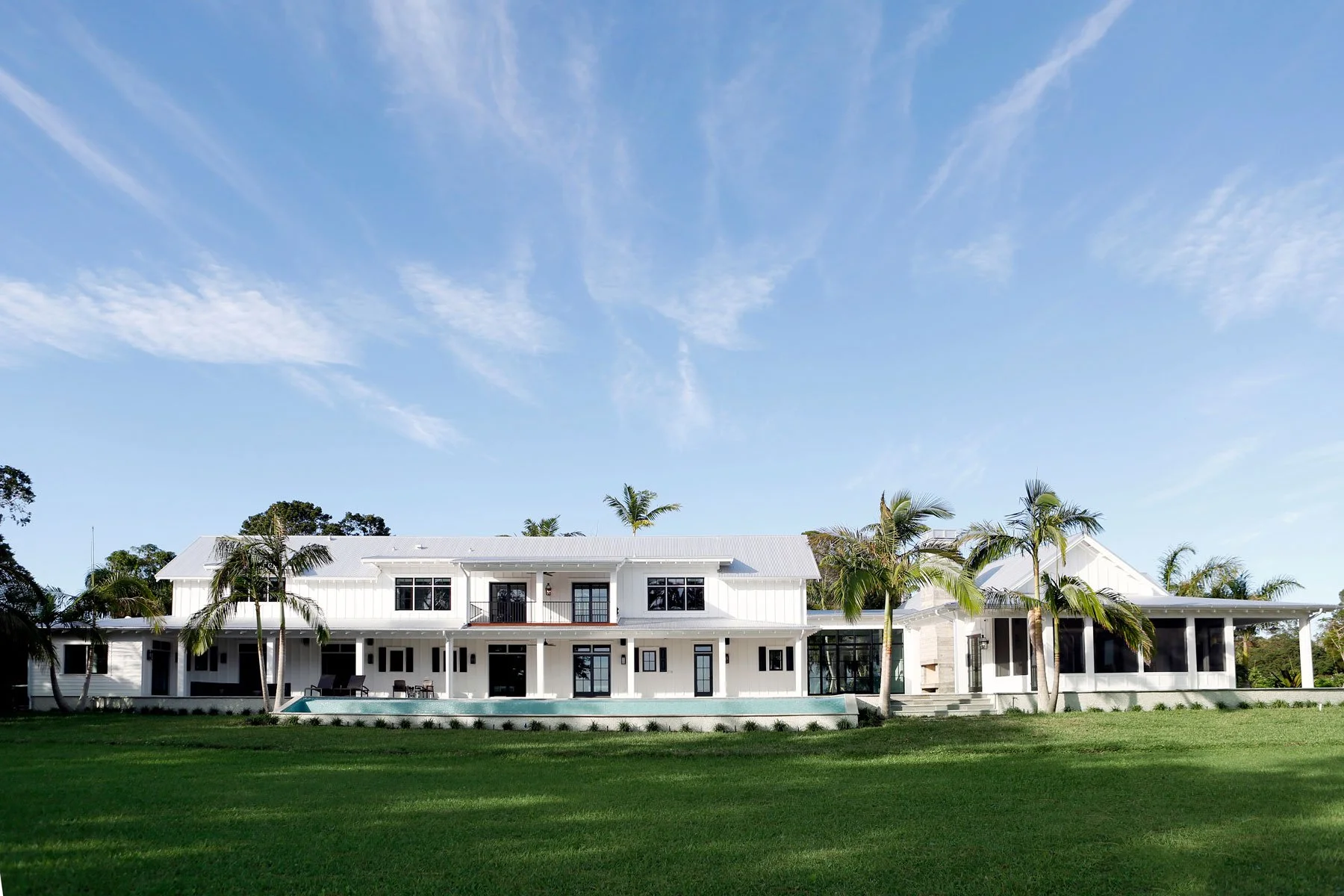 A large white modern house with a wrap-around porch, surrounded by palm trees and a well-maintained lawn, under a blue sky with wispy clouds