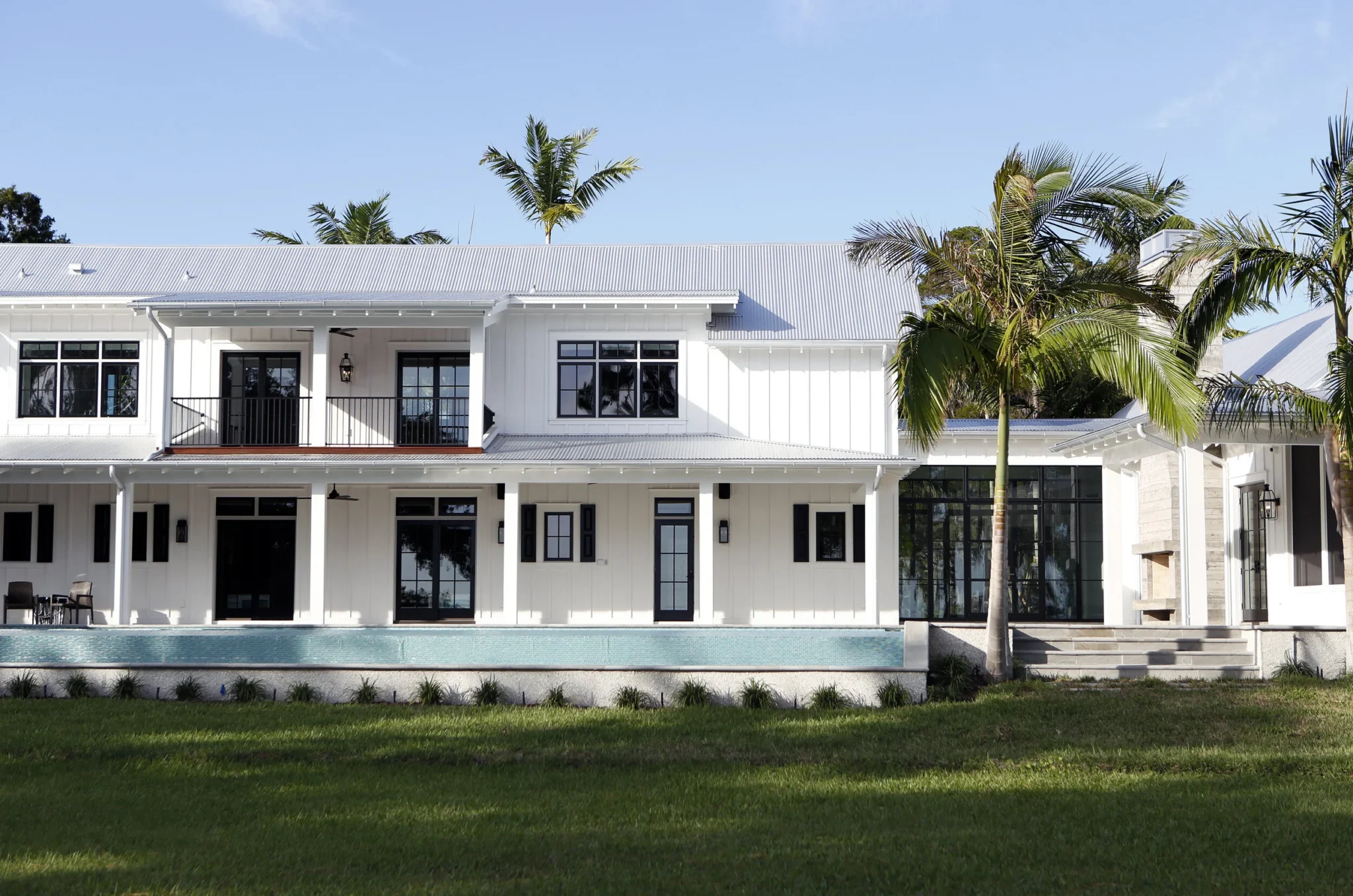 A two-story white house with black window frames, a balcony, and steps leading to the entrance, surrounded by grass and palm trees under a blue sky