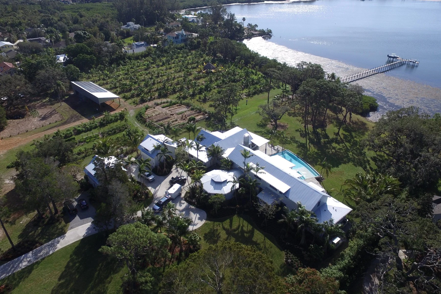 A large house with white metal roofs near a waterfront, surrounded by lush trees and a well-maintained yard
