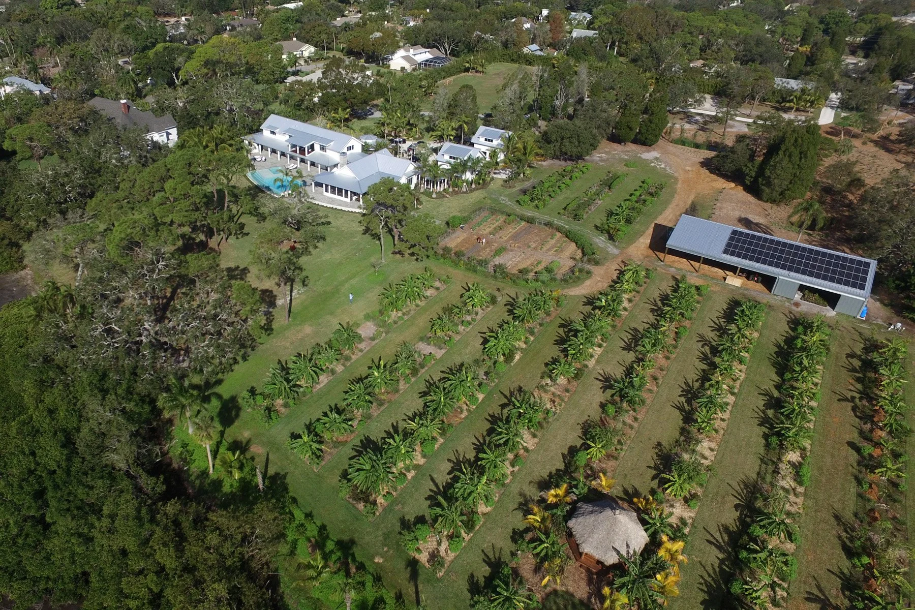 Aerial view of a large rural property with a house, swimming pool, garden plots, and a barn with solar panels, surrounded by trees and greenery