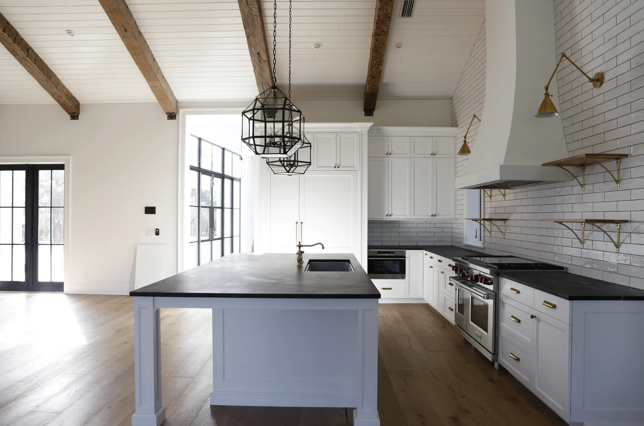 Modern kitchen with white cabinets, black countertops, and wooden ceiling beams, featuring a large island with a sink and gold fixtures, gold shelves