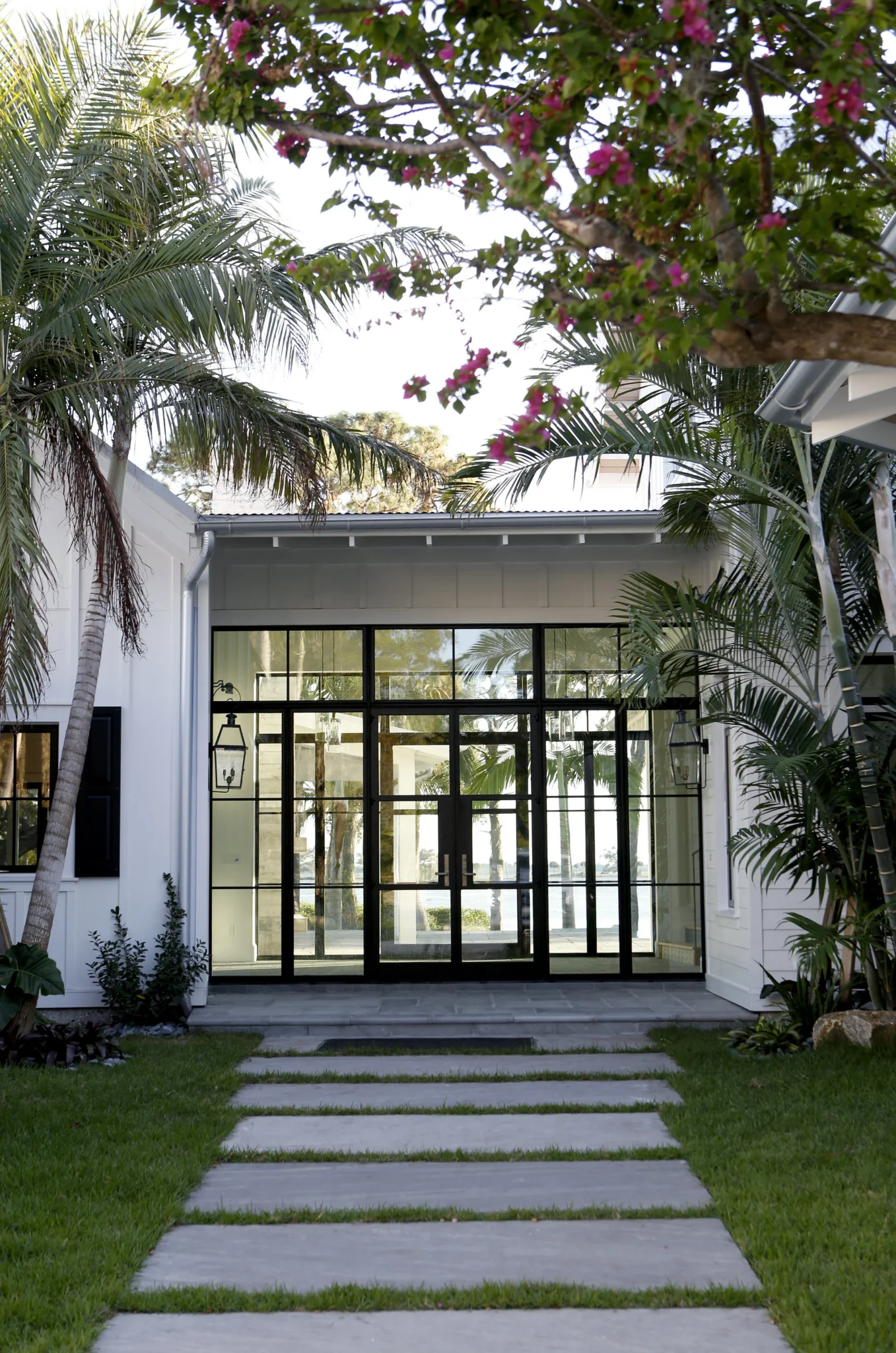 Modern house with glass doors, surrounded by palm trees and flowering plants, with a stone pathway leading to the entrance