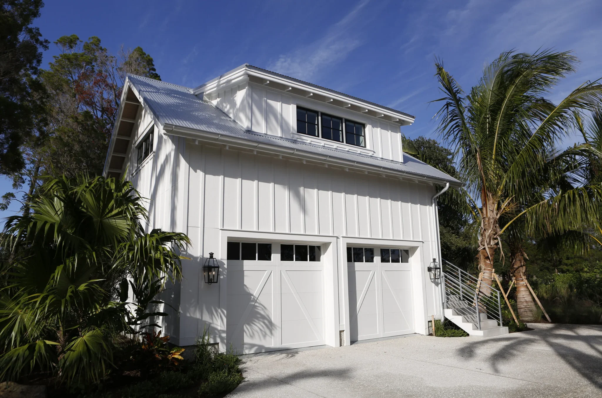 A white modern two-story garage with a metal roof, surrounded by tropical palm trees and plants, against a clear blue sky