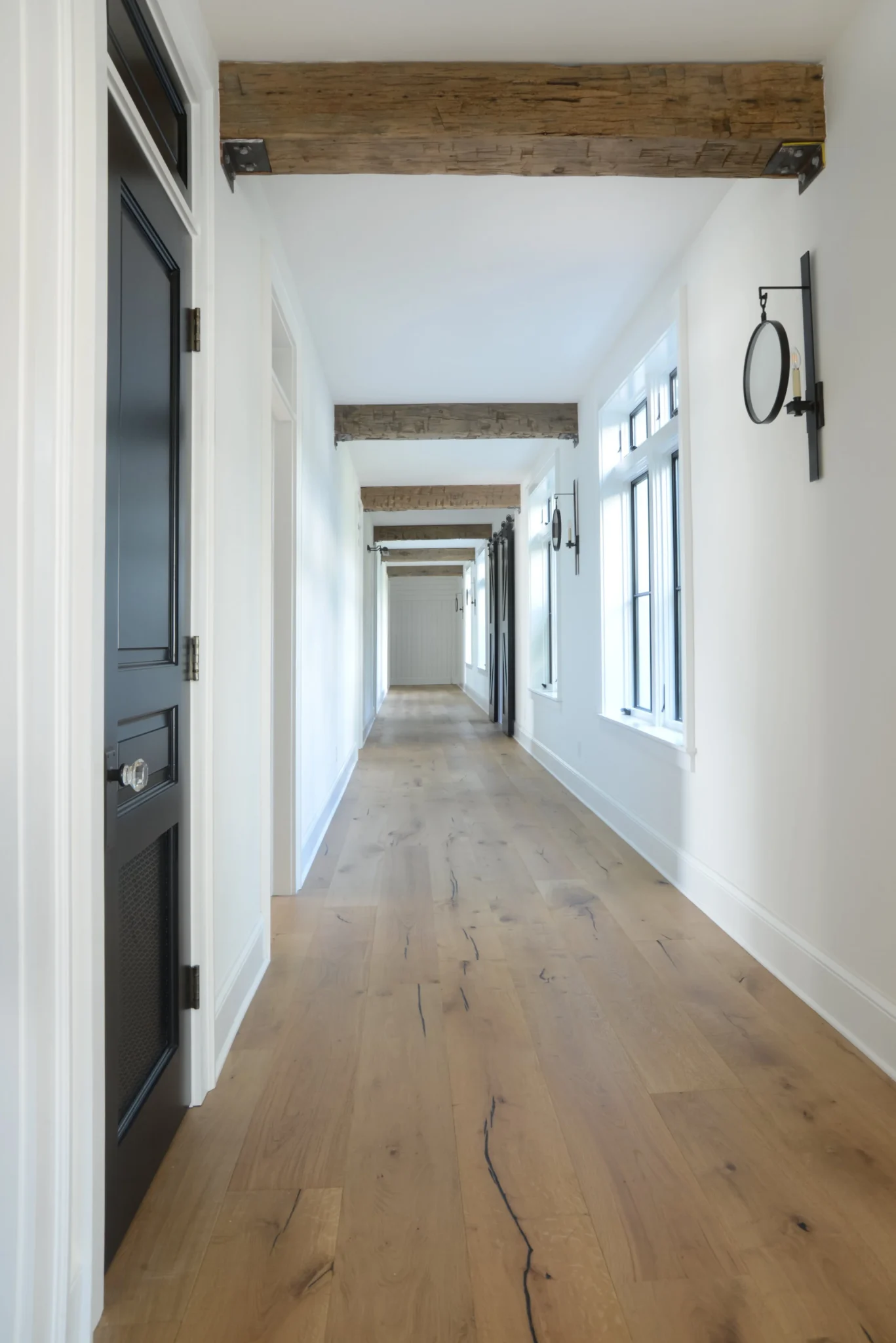 Long hallway with white walls, wooden floors, black doors, windows, and wooden beams