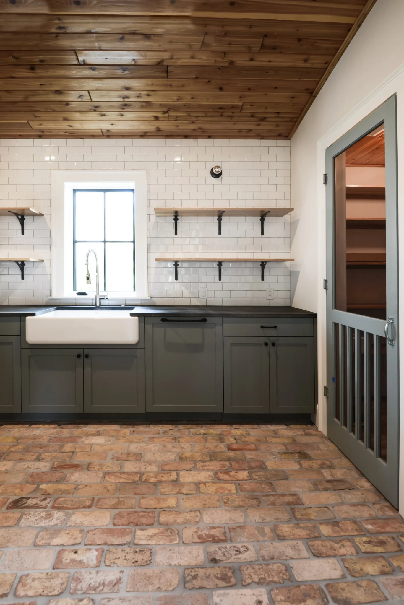 Kitchen with brick floor, gray cabinets, white subway tile backsplash, open shelves, and a window with a farmhouse sink