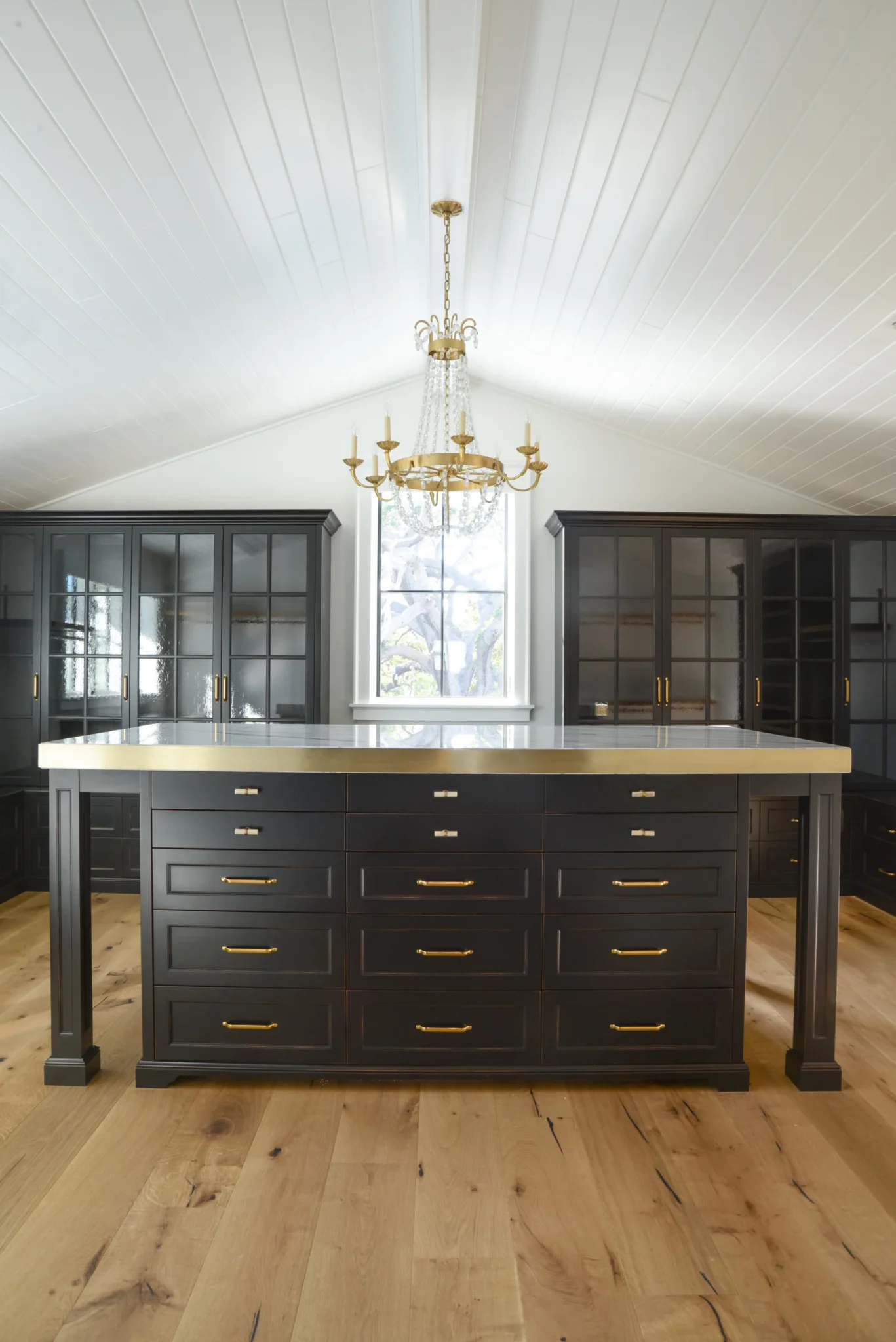 A kitchen with black cabinetry, a central island with a marble top, a window with natural light, a chandelier hanging from the ceiling