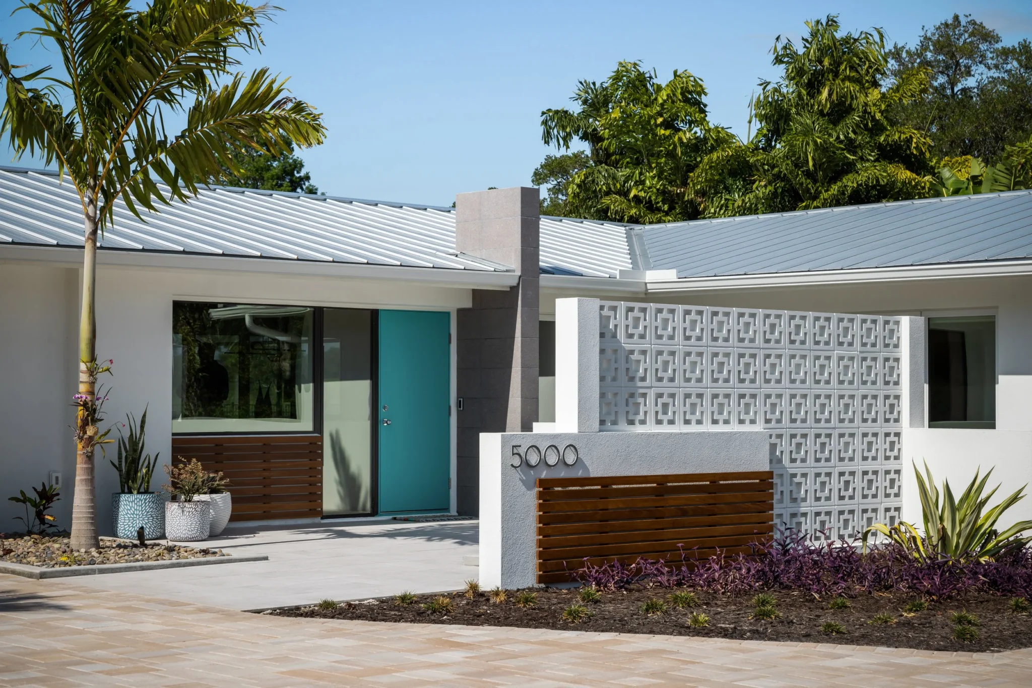 Modern house with a private entrance, turquoise door, decorative concrete block wall, and landscaped front yard with potted plants and trees