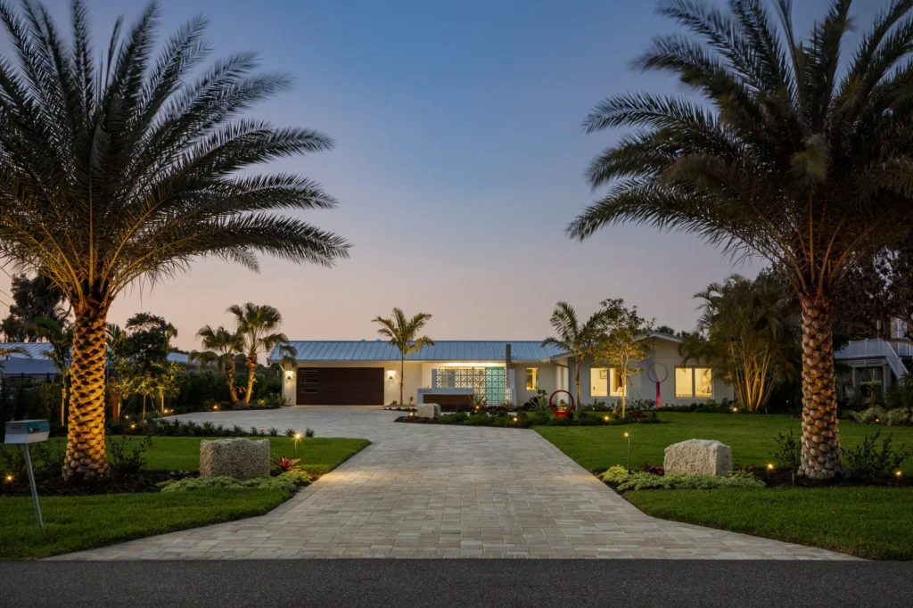 A modern house with a paved driveway, surrounded by palm trees and landscaped garden, evening outdoor lighting