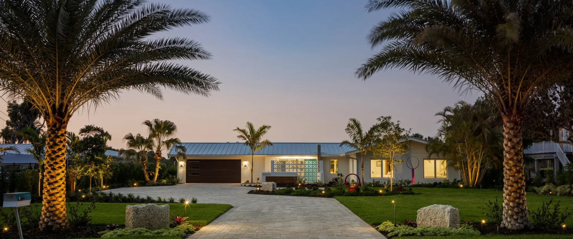 A modern house with a paved driveway, surrounded by palm trees and landscaped garden, evening outdoor lighting
