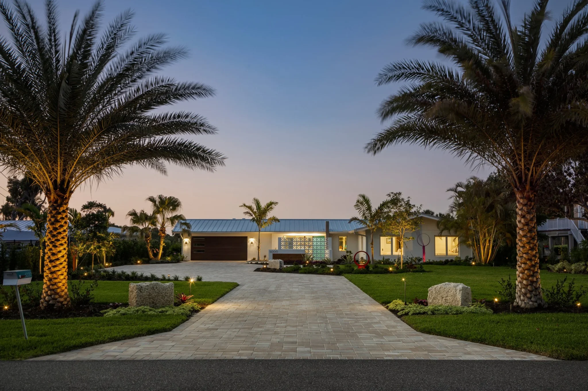 A modern house with a paved driveway, surrounded by palm trees and landscaped garden, evening outdoor lighting