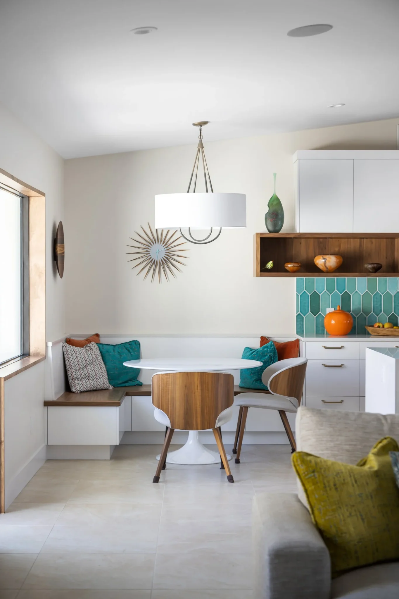 A cozy kitchen nook with a round white table, two wooden and white chairs, built-in corner bench with teal and patterned pillows, window on the left