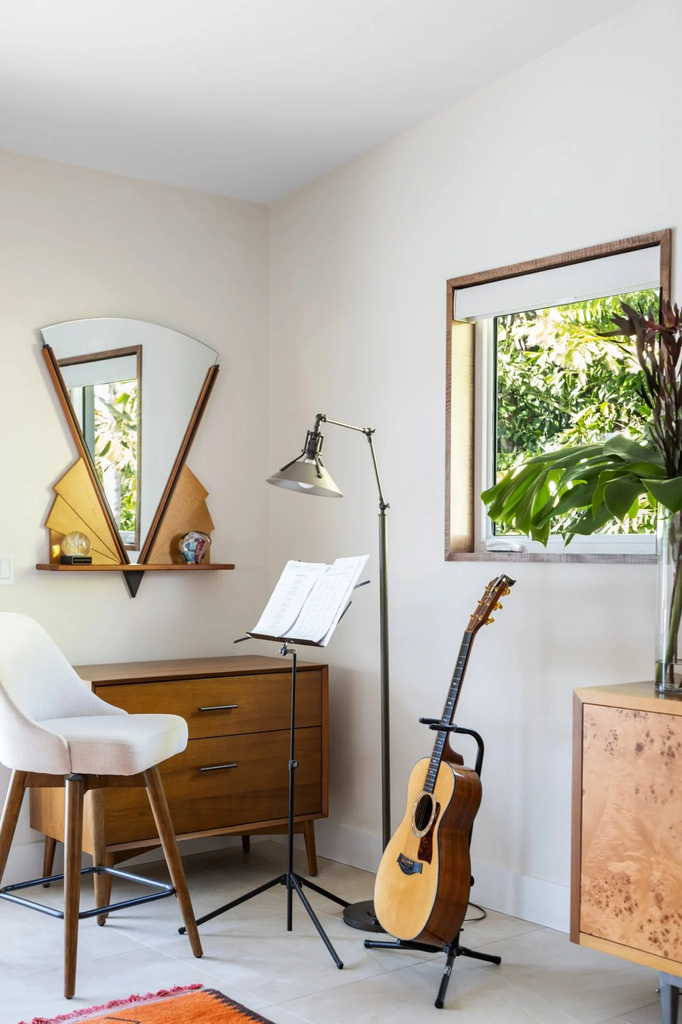 Corner of a room with a guitar on a stand, music stand with sheet music, a white chair, a wooden dresser, a window with lush greenery outside