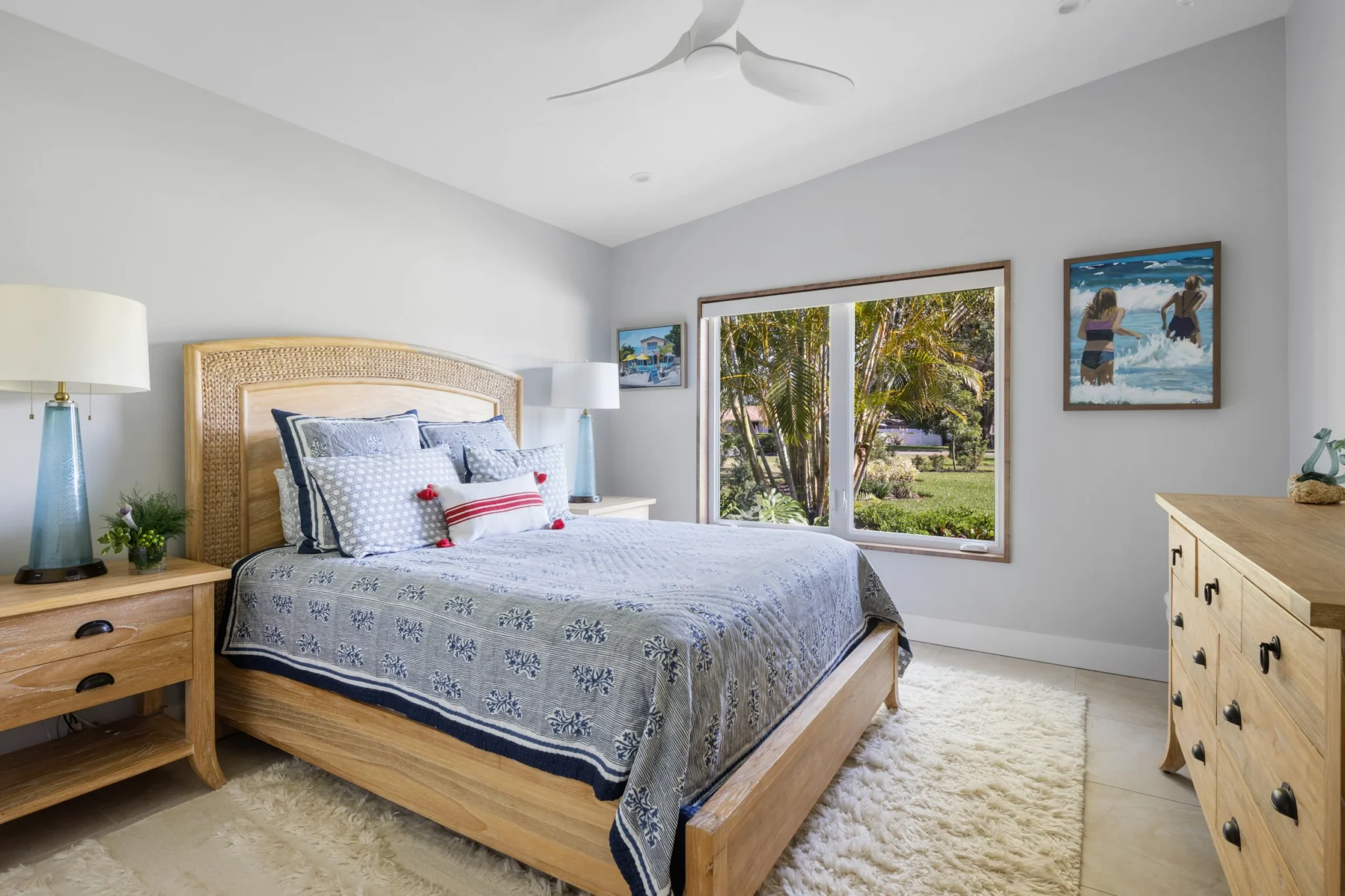 A bedroom with a wooden bed, blue and white bedding, two nightstands with lamps, a wooden dresser, artwork