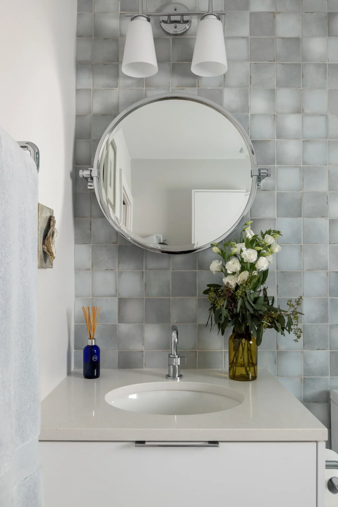 Bathroom sink area with a round mirror, white countertop, a small faucet, a blue vase with reed sticks, and a larger brown vase with white flowers