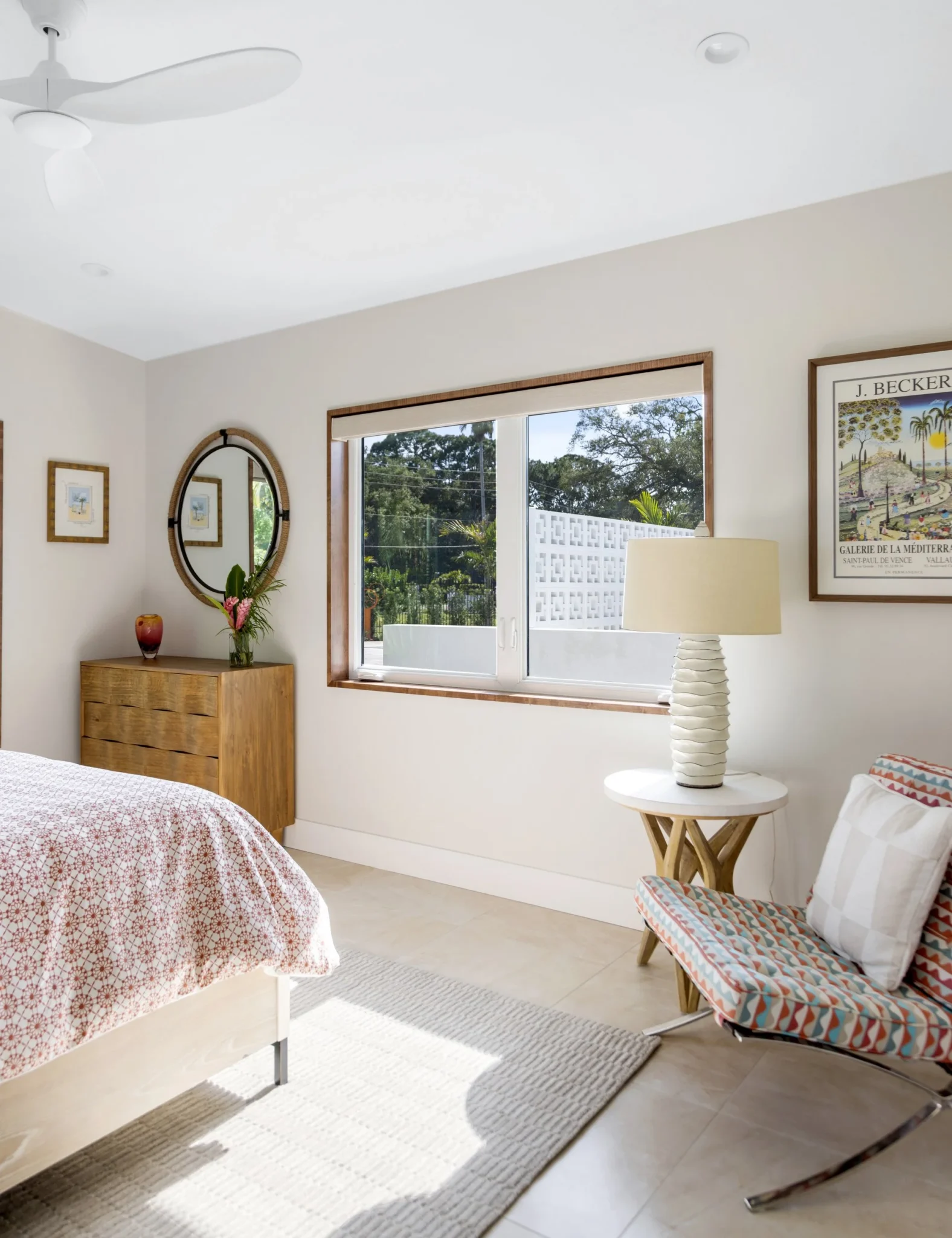 Bright bedroom with a bed, a wooden dresser with a mirror and flowers, a window showing greenery, a modern table lamp