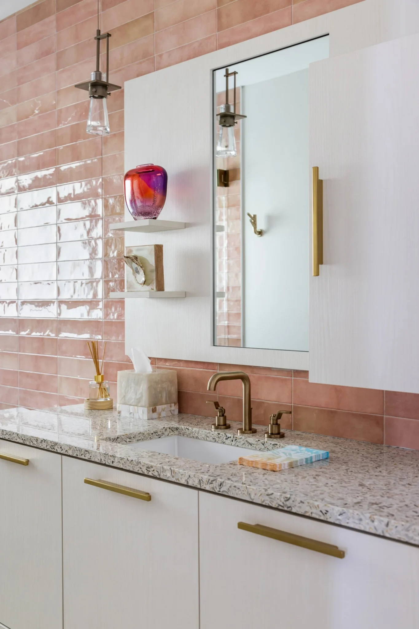 Bathroom vanity with pink tiled wall, white cabinets with gold handles, granite countertop, and brass faucet