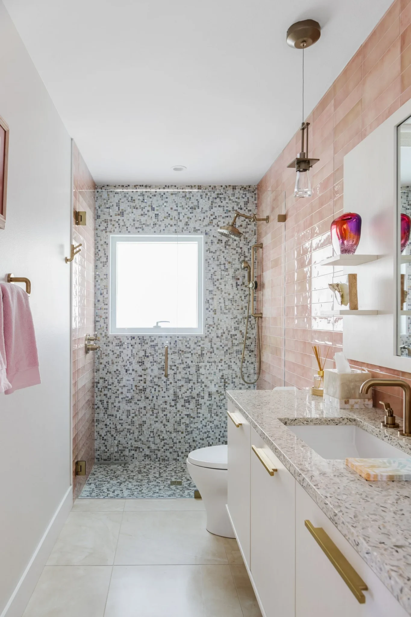 Modern bathroom with walk-in shower, pink tiles, mosaic wall, white vanity with granite countertop, pink towels, decorative vases, and large window