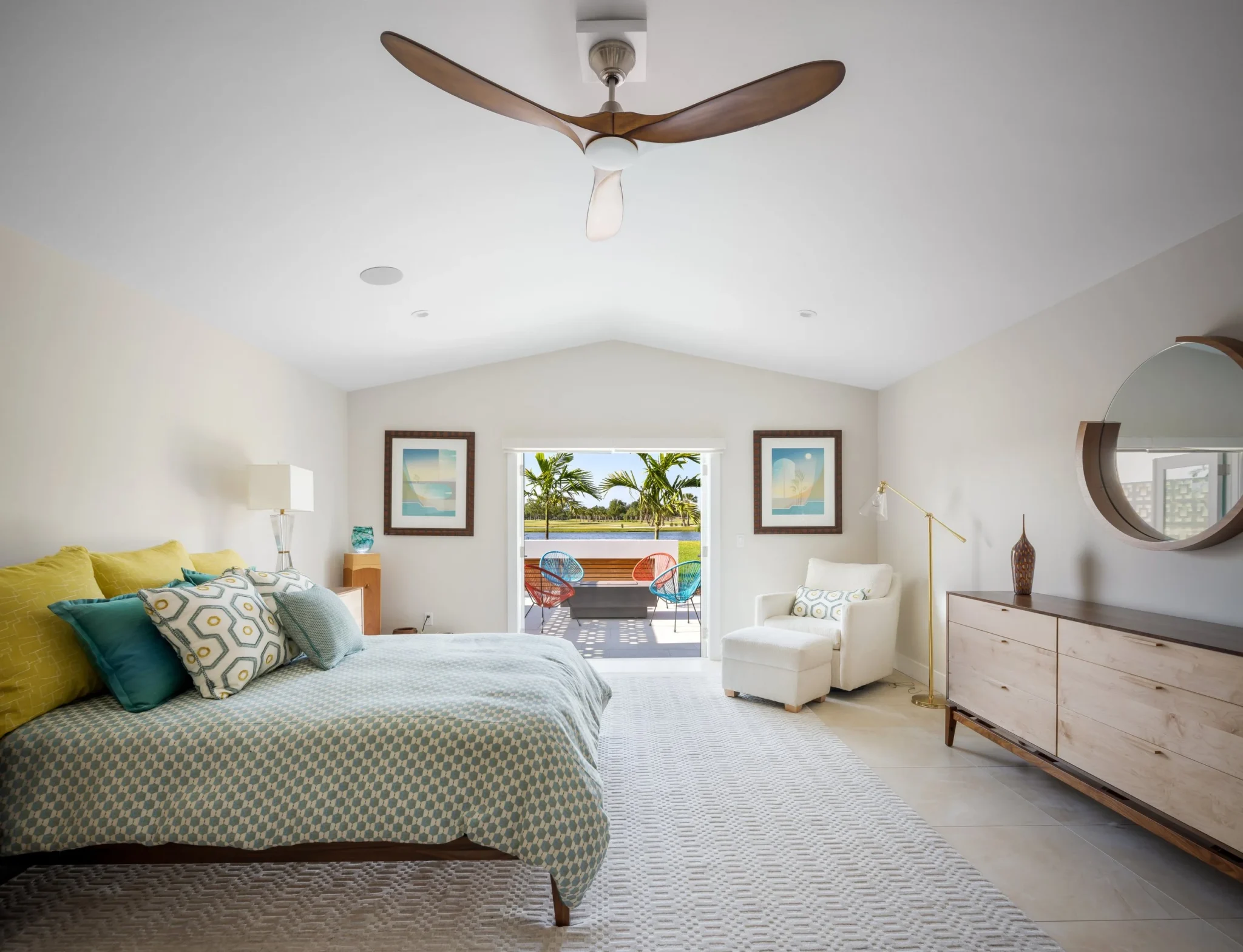 A bedroom with a large bed featuring patterned bedspread and pillows, a white armchair with a pillow, a wooden sideboard, a mirror
