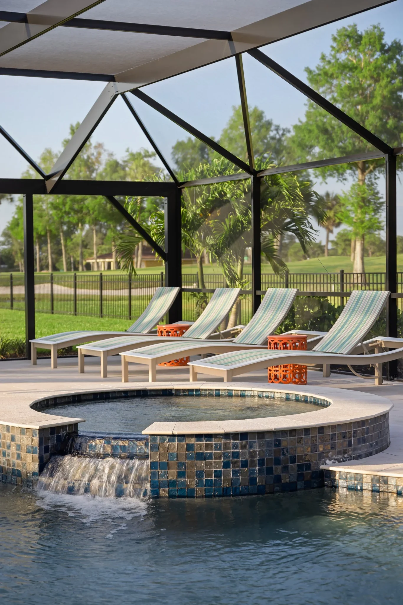 Four pool loungers with striped cushions and orange side tables on a poolside patio, with a small waterfall feature and screened enclosure