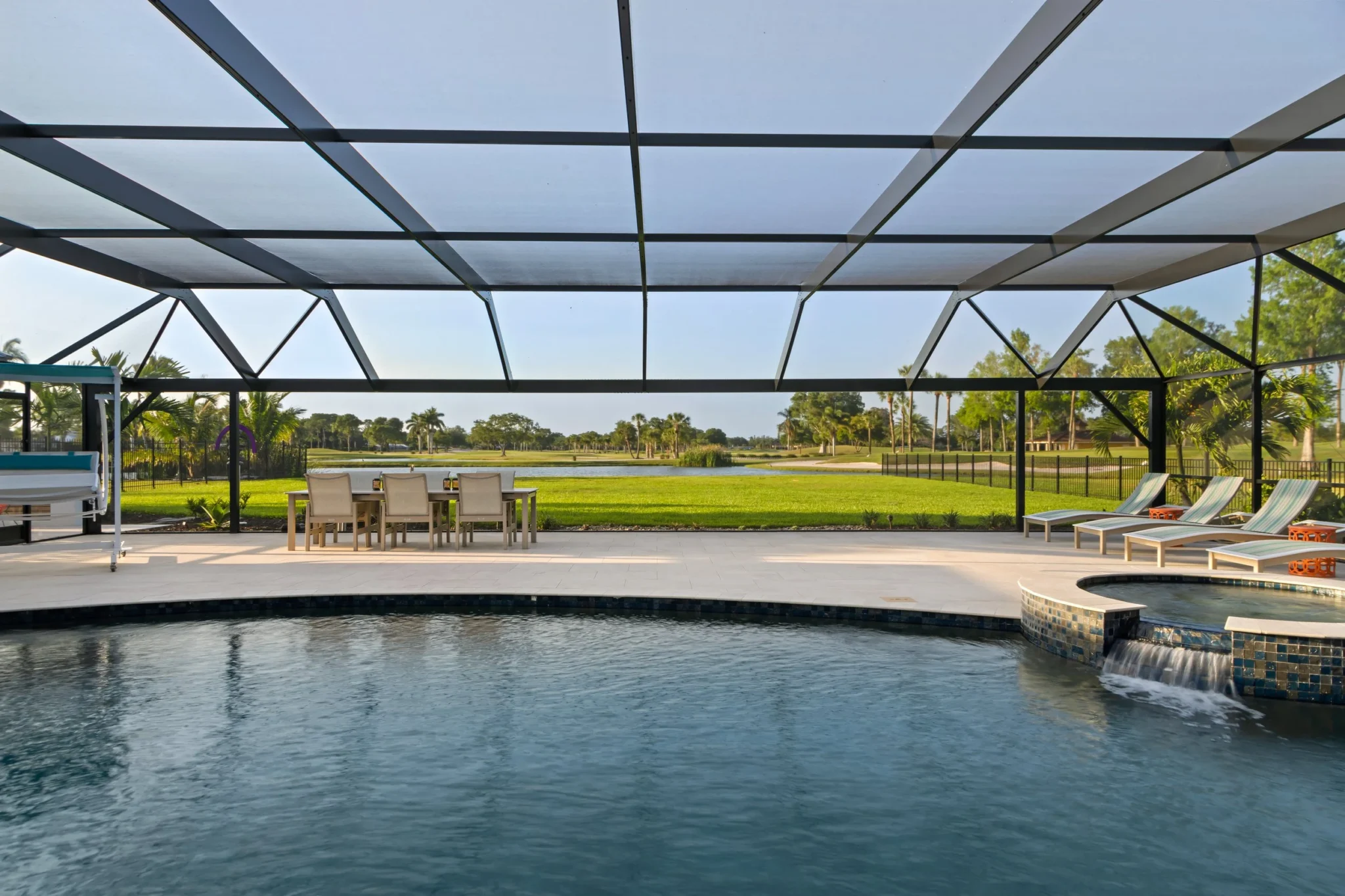 Backyard view of a swimming pool with a screened enclosure, lounge chairs, a dining table with chairs, and a grassy yard with trees in the distance