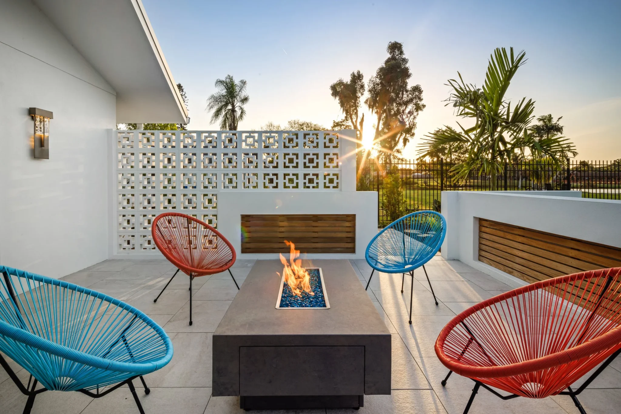 Outdoor patio with colorful circular chairs around a fire pit, white walls, lattice fence, palm trees, and sunset in the background