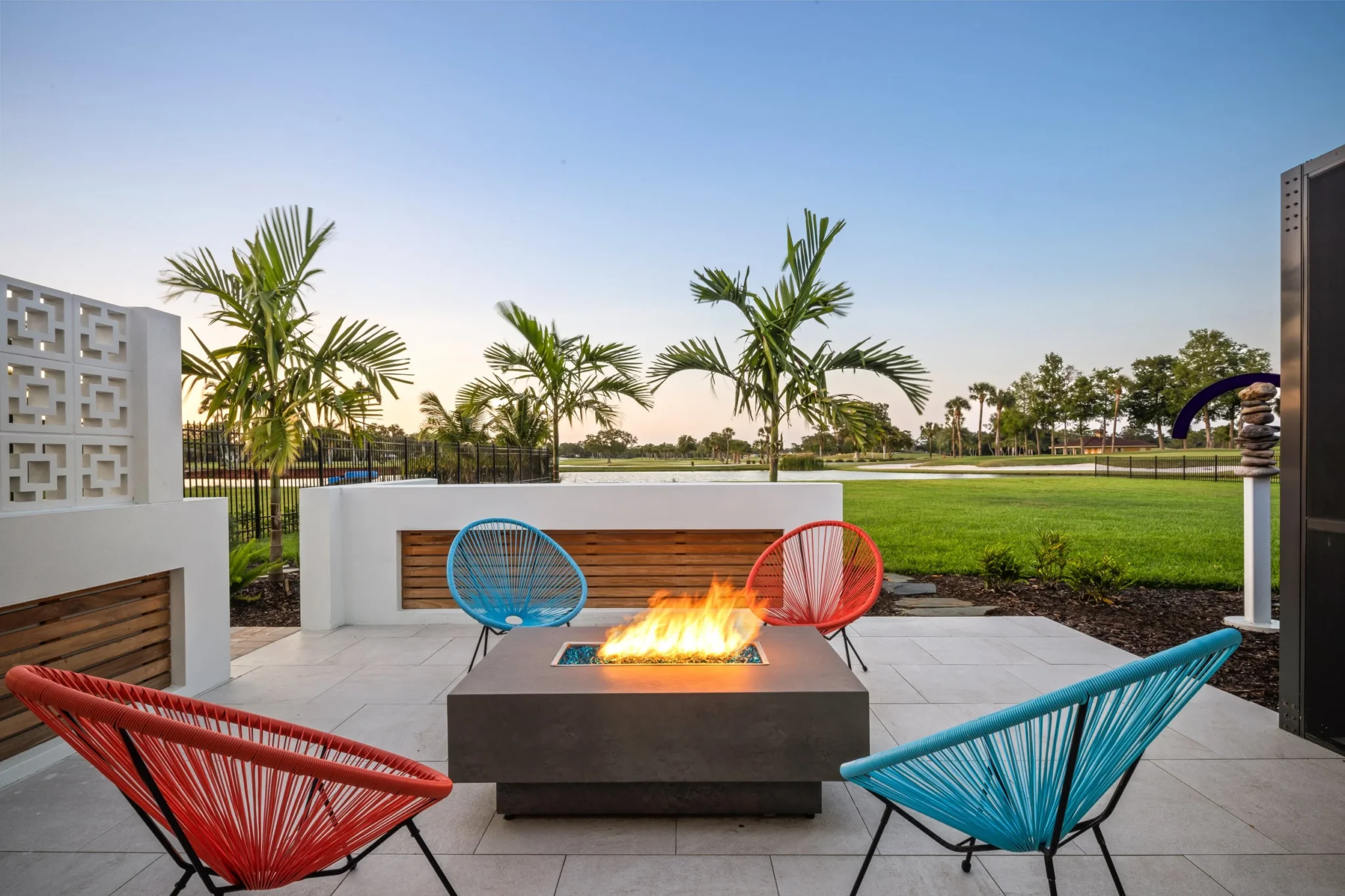 Outdoor patio with four colorful chairs around a fire pit, palm trees, green grass, and a lake in the background
