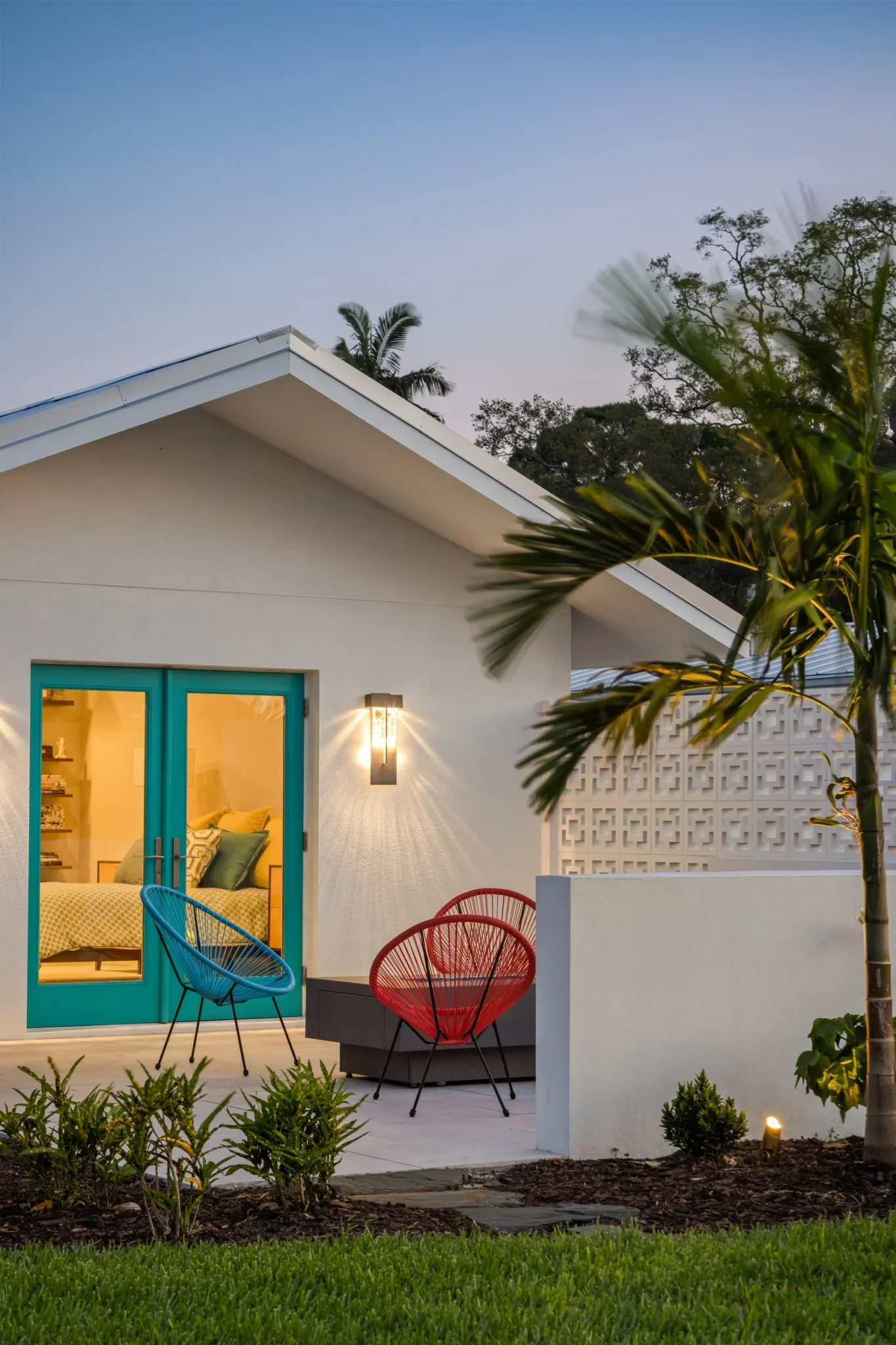 Exterior view of a modern house with a white facade and turquoise double doors, illuminated by outdoor lighting, with colorful chairs on the patio